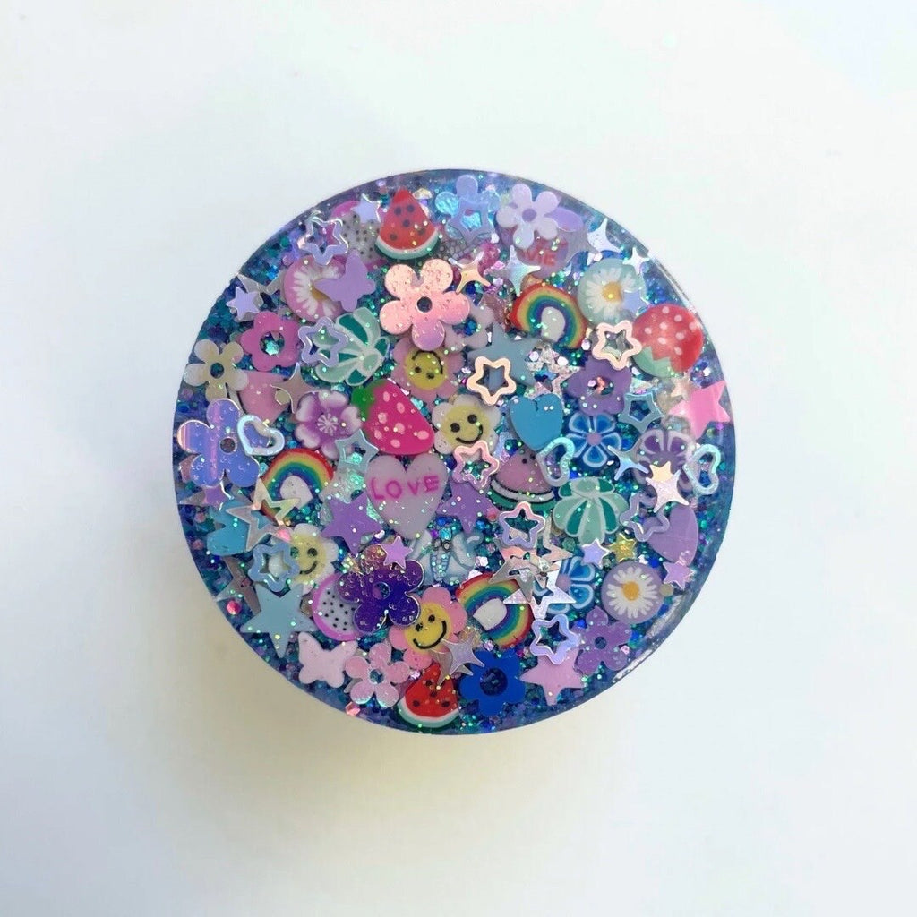 
A circular phone grip featuring a maximalist, multi-colored design on a dark blue glitter base. The surface is densely packed with 3D and flat charms, including:

Fruit and Icons: Strawberries, watermelon slices, rainbows, smiley faces, and a purple heart with the word "LOVE".

Decorative Elements: Pastel flowers, butterflies, and numerous shimmering holographic stars in various sizes.

Finish: The design is encased in a high-gloss, clear resin dome and is shown against a plain white background.