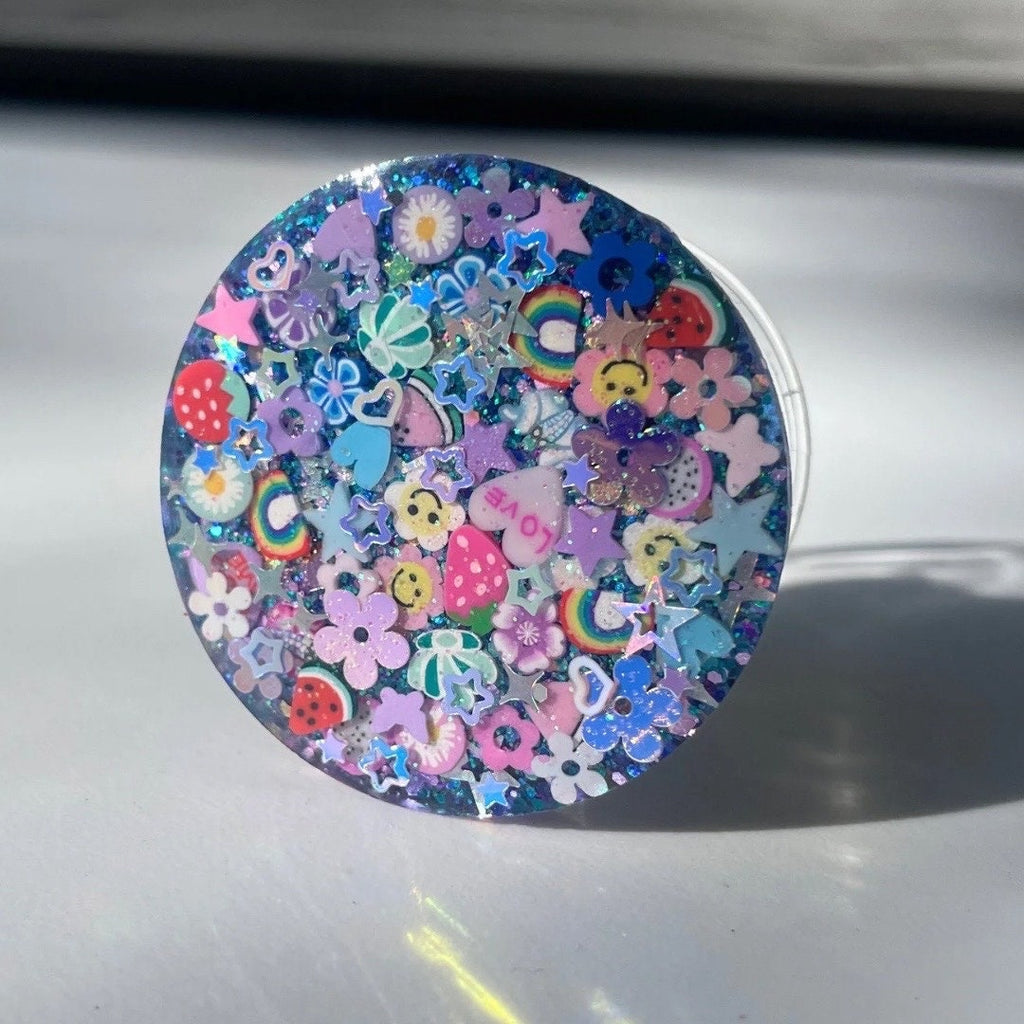 
A circular phone grip featuring a maximalist, multi-colored design on a dark blue glitter base. The surface is densely packed with 3D and flat charms, including:

Fruit and Icons: Strawberries, watermelon slices, rainbows, smiley faces, and a purple heart with the word "LOVE".

Decorative Elements: Pastel flowers, butterflies, and numerous shimmering holographic stars in various sizes.

Finish: The design is encased in a high-gloss, clear resin dome and is shown against a plain white background.