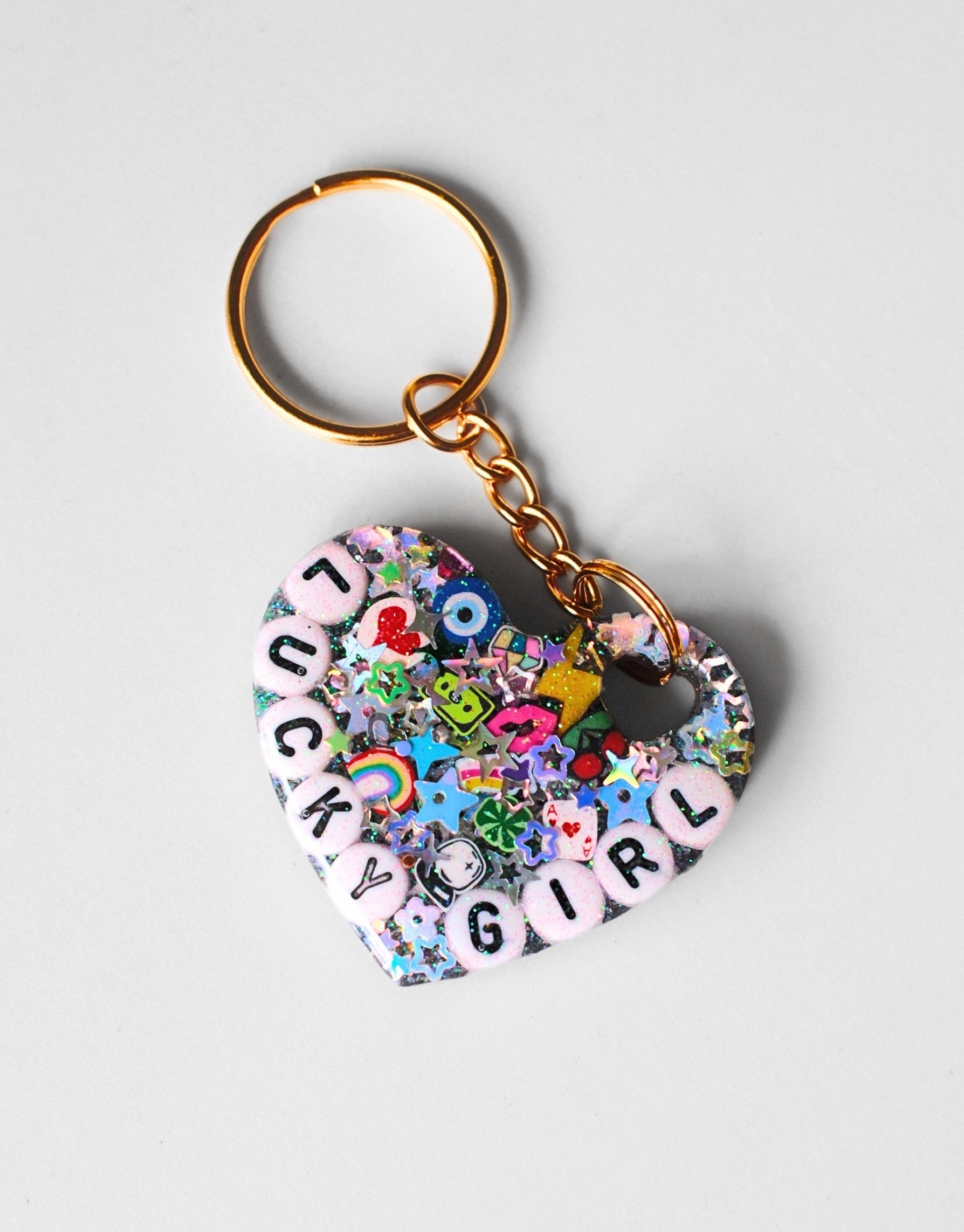 A heart-shaped keychain featuring a colorful collage of stickers, charms, and pink letter tiles spelling 'JACKY GURL' on a light gray background.