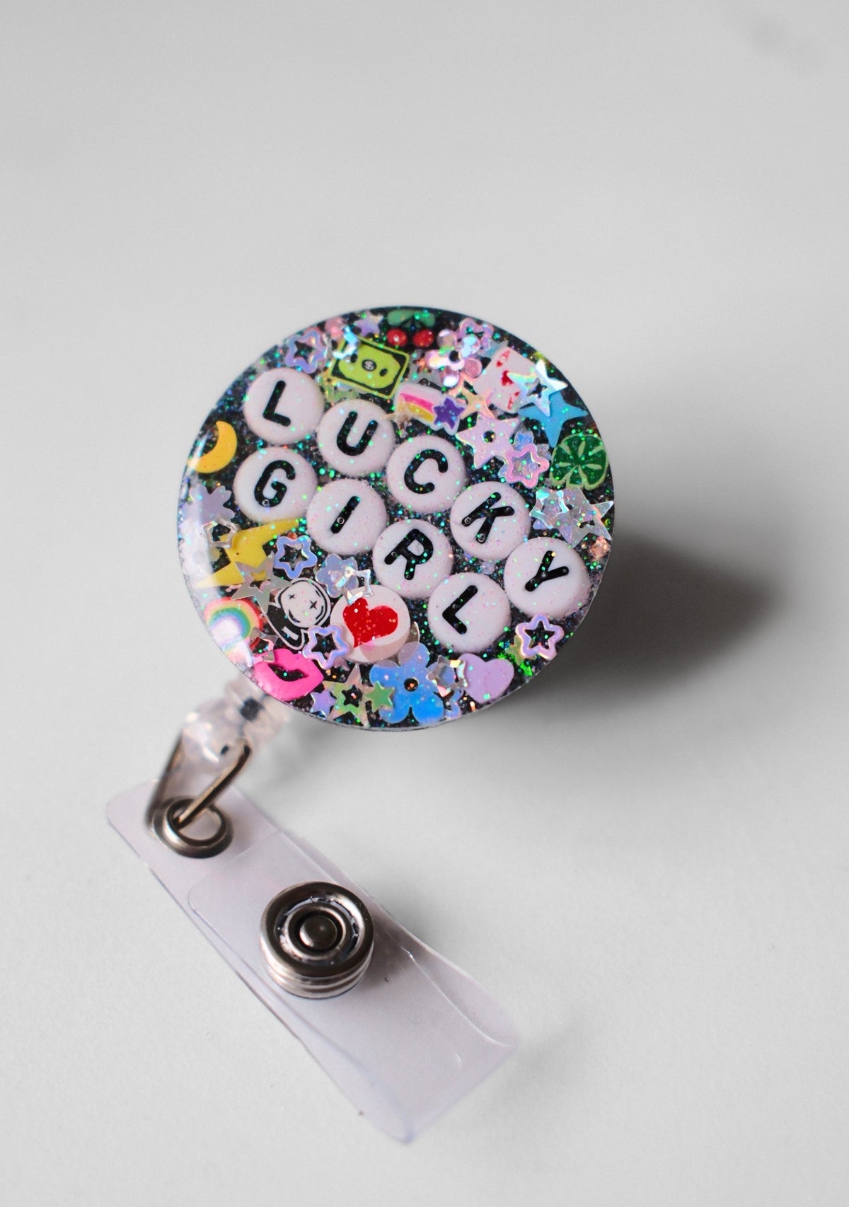 A round badge reel cover featuring a colorful 'LUCKY GIRL' letter tile design over a multicolor glitter and floral background, attached to a clear plastic clip with a metal snap button, shown on a light gray background.