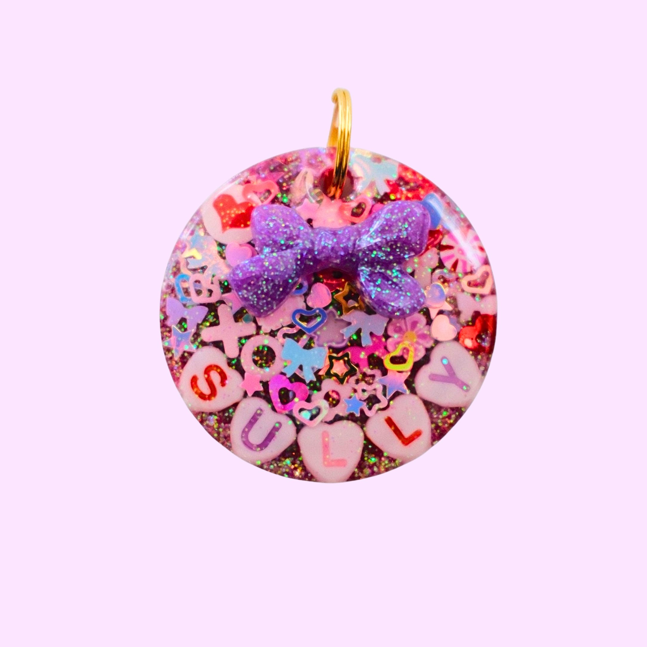 Circle-shaped pink and red Valentine-themed pet ID tag decorated with heart-shaped letter beads spelling the pet’s name and a 3D purple bow charm