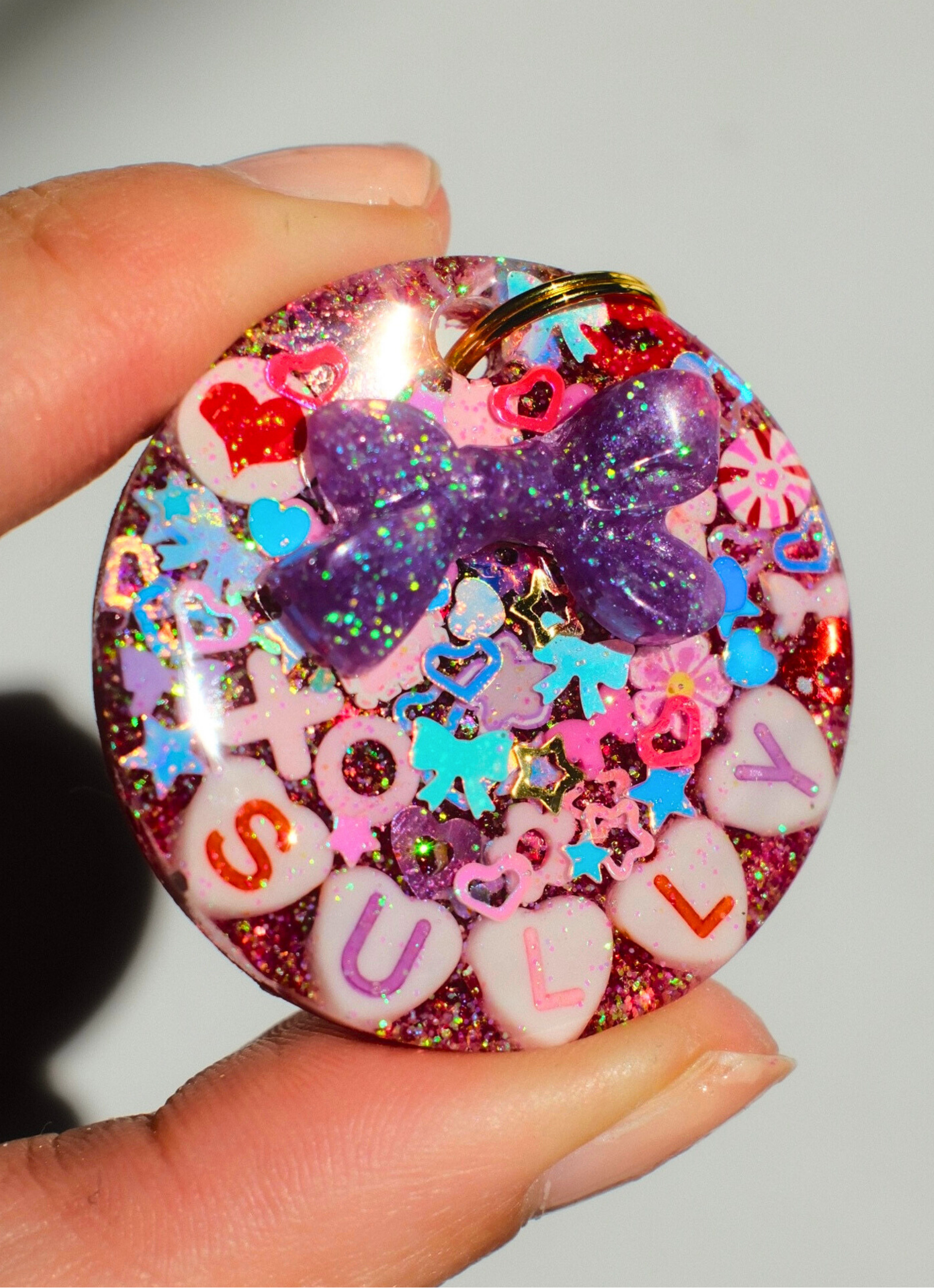 Circle-shaped pink and red Valentine-themed pet ID tag decorated with heart-shaped letter beads spelling the pet’s name and a 3D purple bow charm held up in the sun