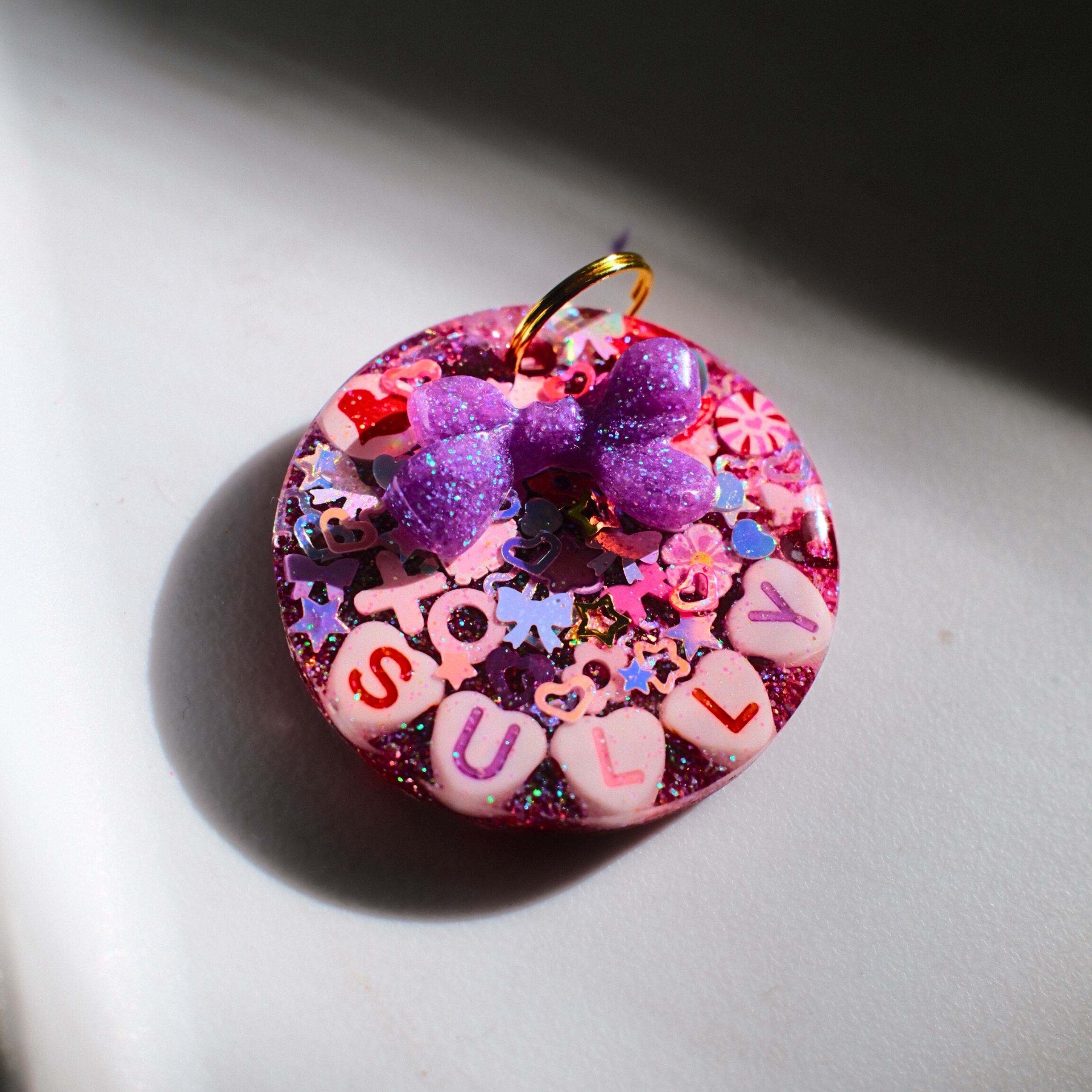 Circle-shaped pink and red Valentine-themed pet ID tag decorated with heart-shaped letter beads spelling the pet’s name and a 3D purple bow charm
