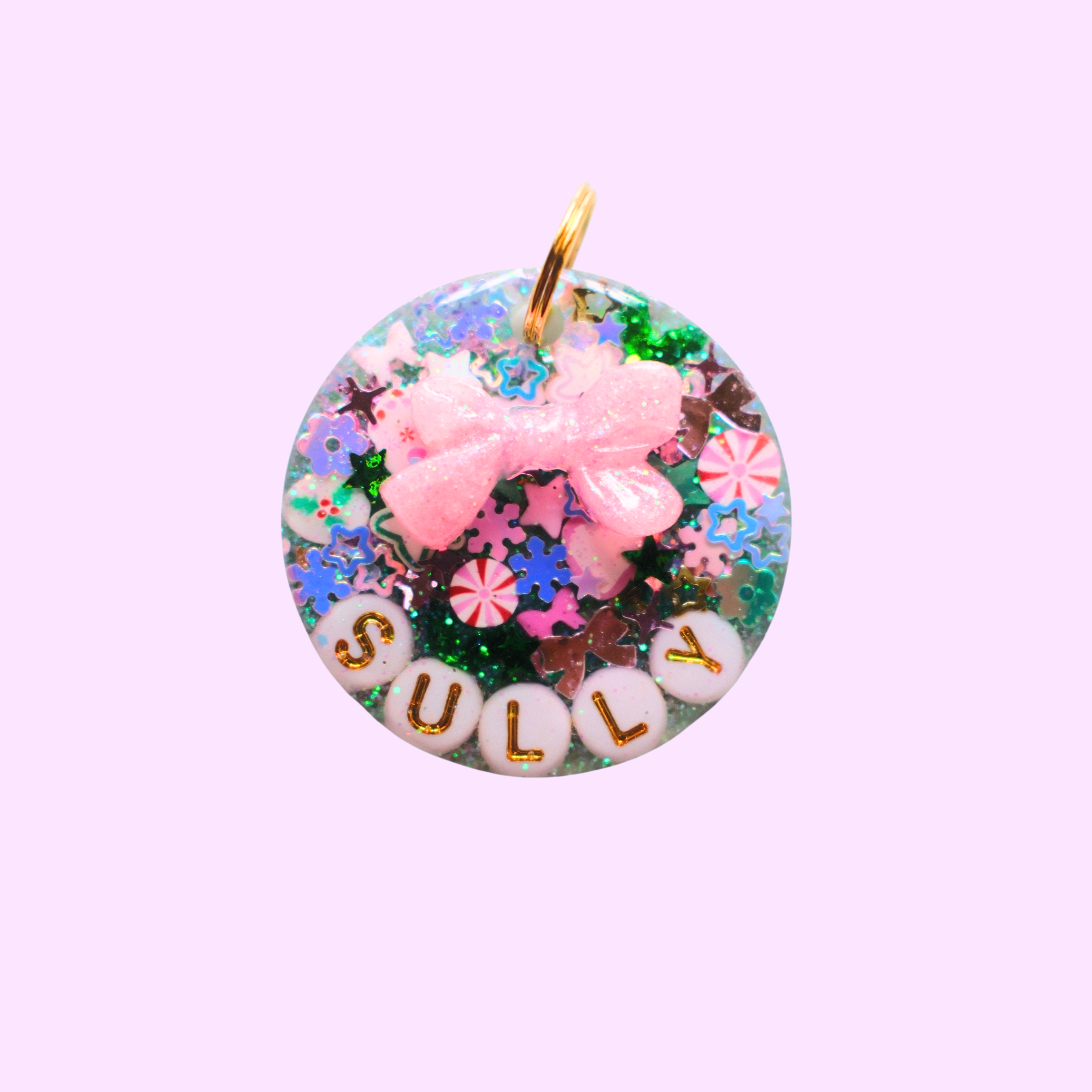 Round resin pet ID tag filled with colorful glitter, star and candy-shaped sprinkles, white letter beads spelling ‘SULLY,’ and a raised pink bow charm, attached to a gold ring