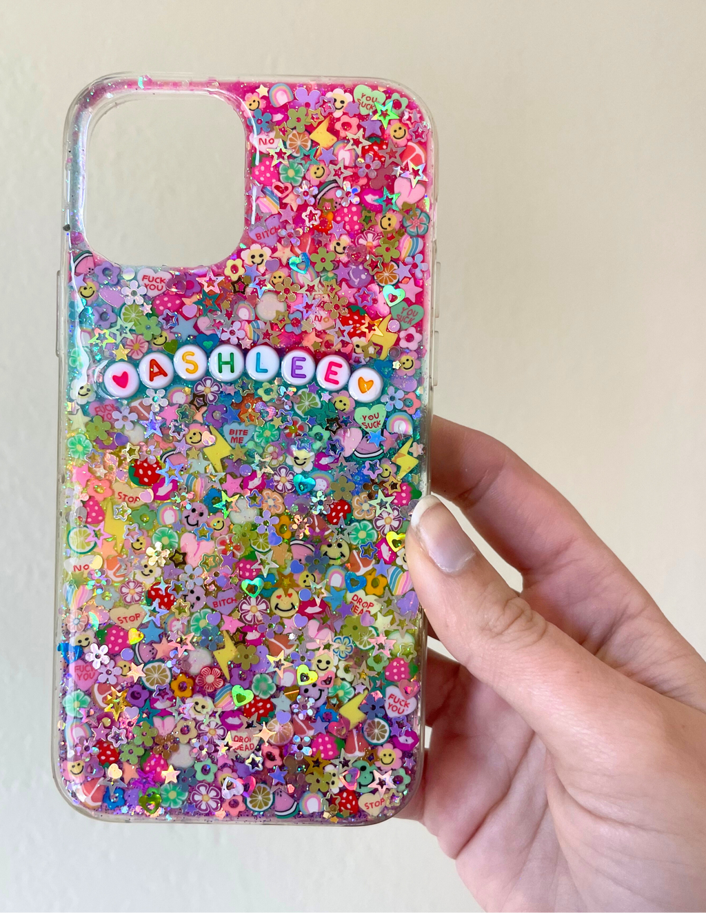 Colorful resin-style phone case filled with multicolored glitter, flowers, stars, smiley faces, fruit and novelty polymer clay slices, featuring white round letter beads spelling ‘ASHLEE’ with heart accents and a hand holding it against a white wall
