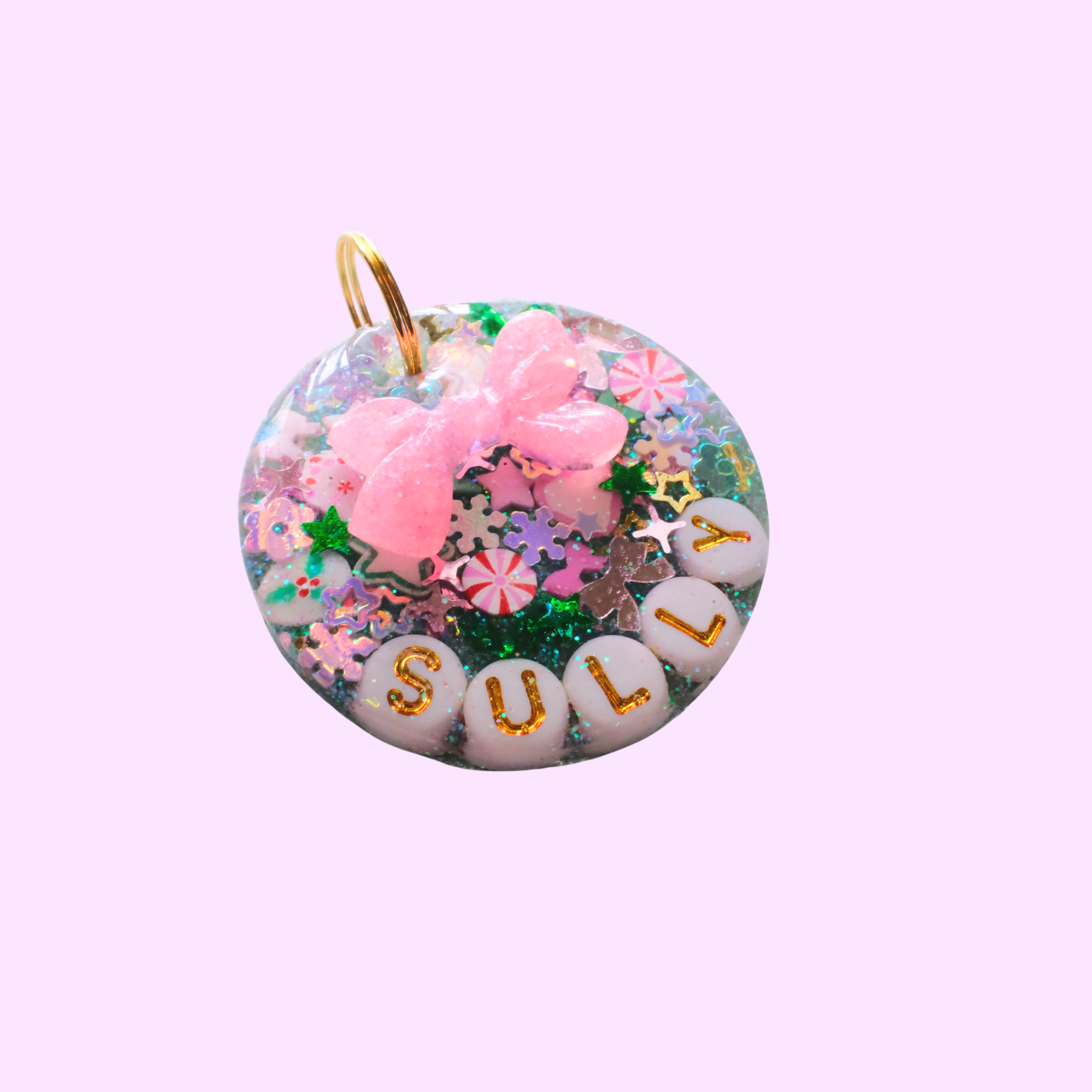 Round resin pet ID tag filled with colorful glitter, star and candy-shaped sprinkles, white letter beads spelling ‘SULLY,’ and a raised pink bow charm, attached to a gold ring