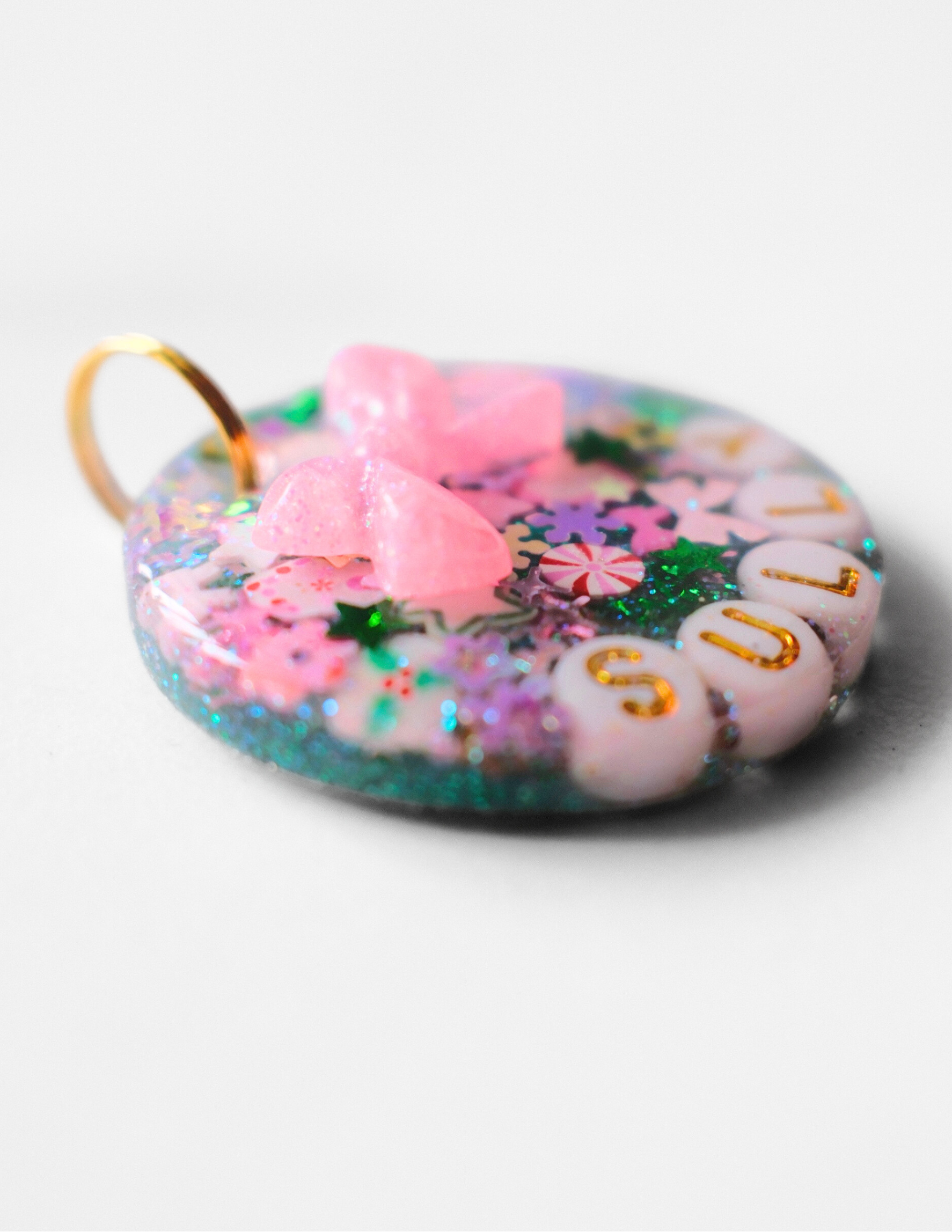 Round resin pet ID tag filled with colorful glitter, star and candy-shaped sprinkles, white letter beads spelling ‘SULLY,’ and a raised pink bow charm, attached to a gold ring