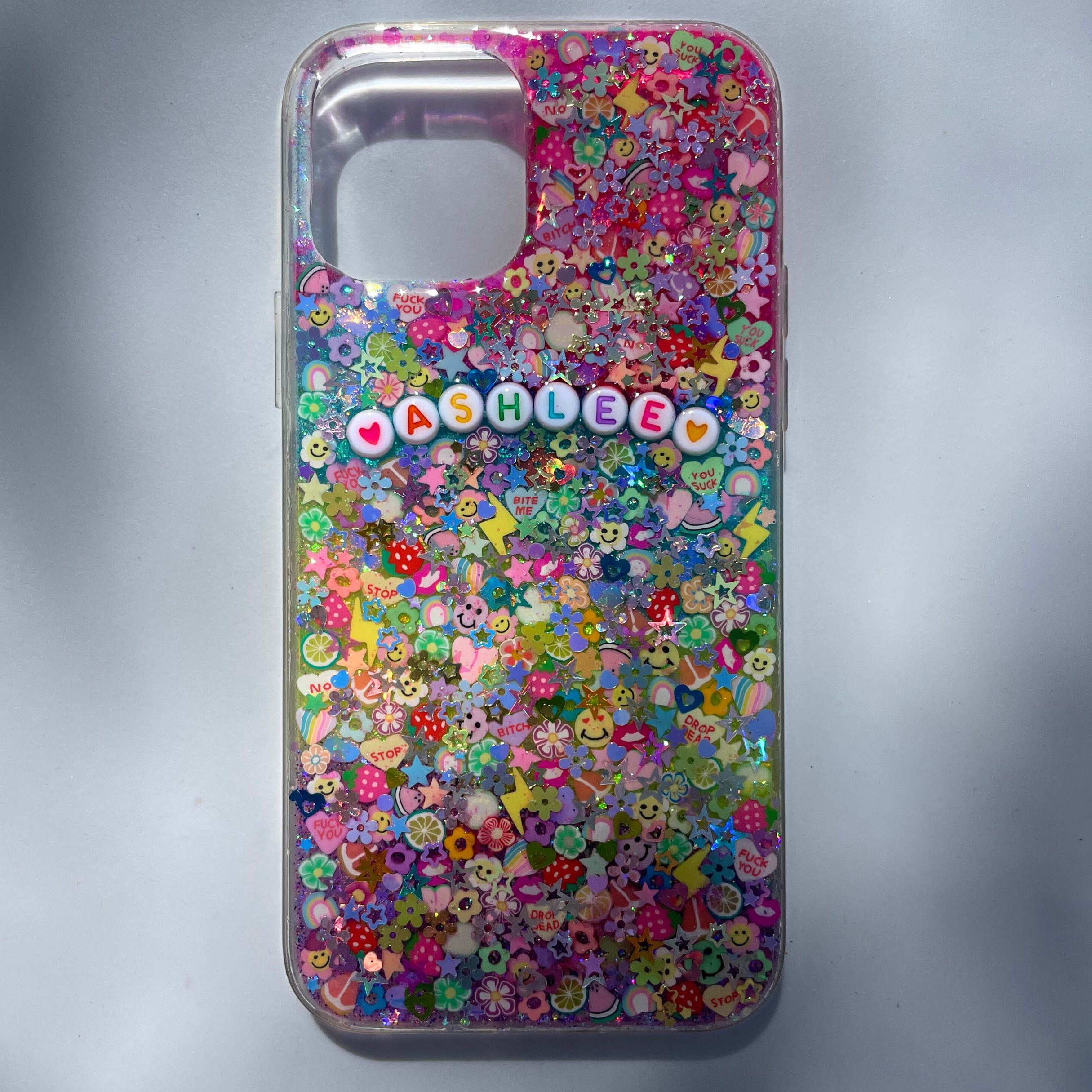 Colorful resin-style phone case filled with multicolored glitter, flowers, stars, smiley faces, fruit and novelty polymer clay slices, featuring white round letter beads spelling ‘ASHLEE’ with heart accents
