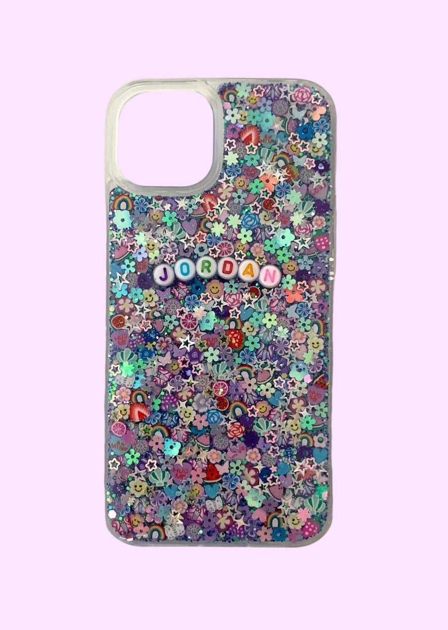 
A clear smartphone case featuring a maximalist, multi-colored design on a purple and teal glitter base. The back is densely packed with 3D charms, including strawberries, rainbow arches, flowers, and holographic stars. White circular beads with multicolored letters are centered on the case, spelling out the name "JORDAN." The case is shown against a soft lavender background with a large rectangular camera cutout at the top.