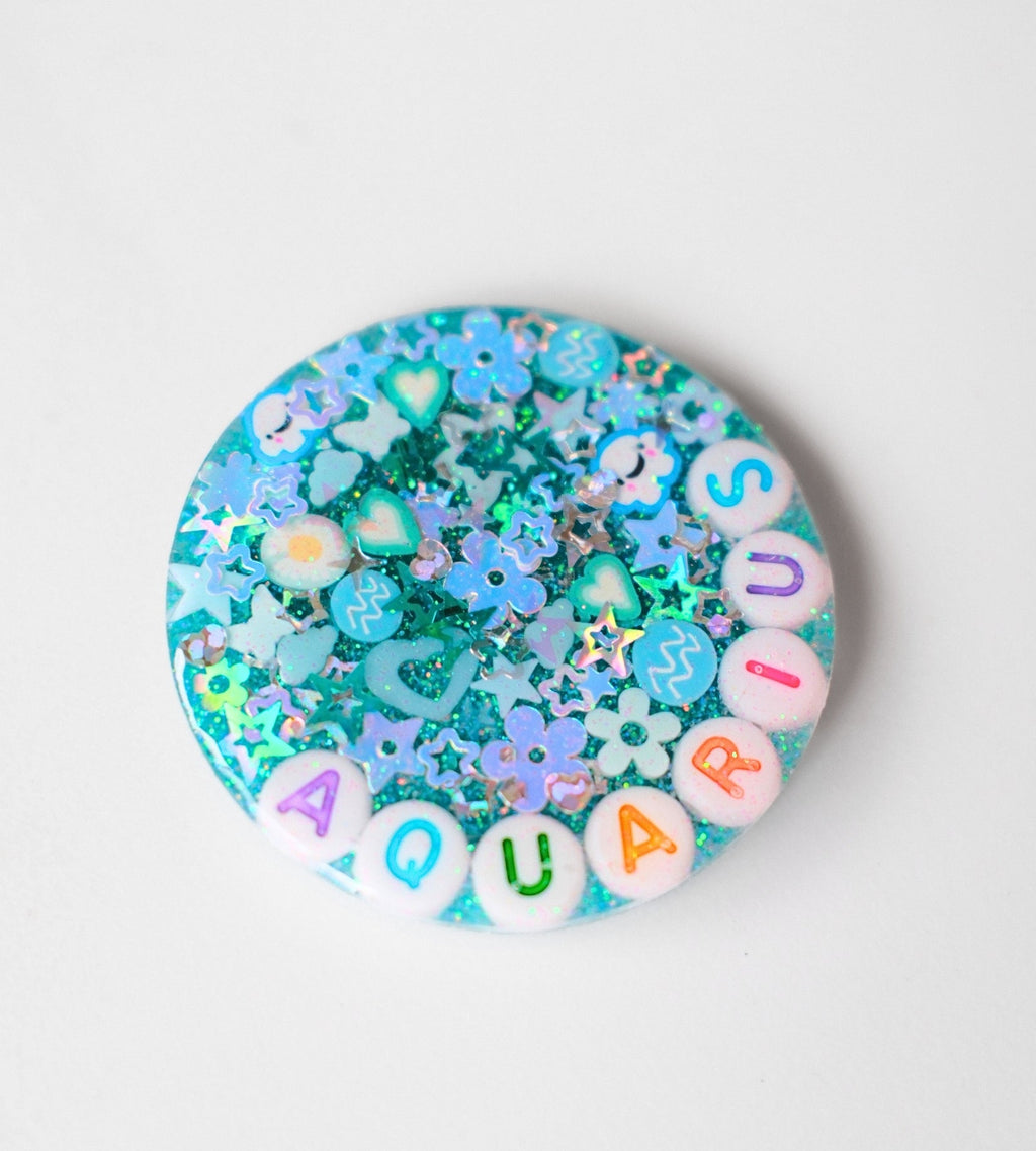 A round decorative item filled with pastel blue and turquoise glitter, star and heart-shaped charms, and white letter beads spelling 'AQUARIUS' on a plain light background.