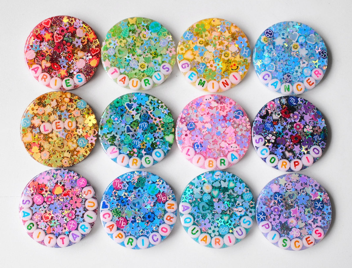 Twelve round resin name badges arranged in a 3x4 grid on a white background. Each badge contains colorful glitter, flower-shaped charms, and raised letter charms spelling out the name 'JUNGHO'.
