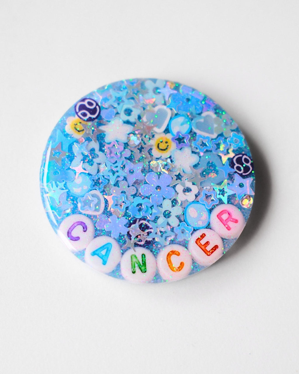 A round blue glitter pinback button featuring the word 'CANCER' spelled out in colorful letter beads, surrounded by blue and purple astrological and zodiac-themed glitter pieces on a light gray background.