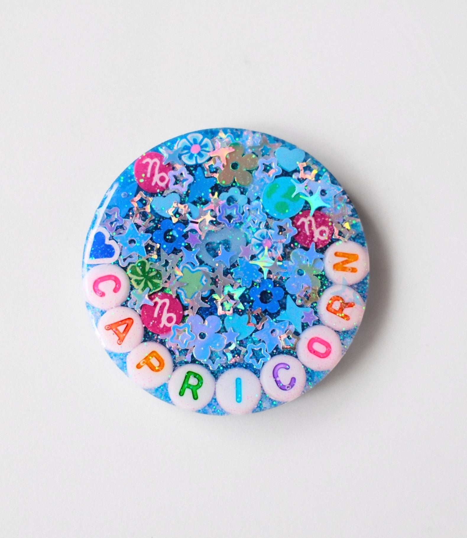 A round blue glittery pinback button featuring the word 'CAPRICORN' spelled out by colorful letter beads, surrounded by blue and silver star-shaped glitter.