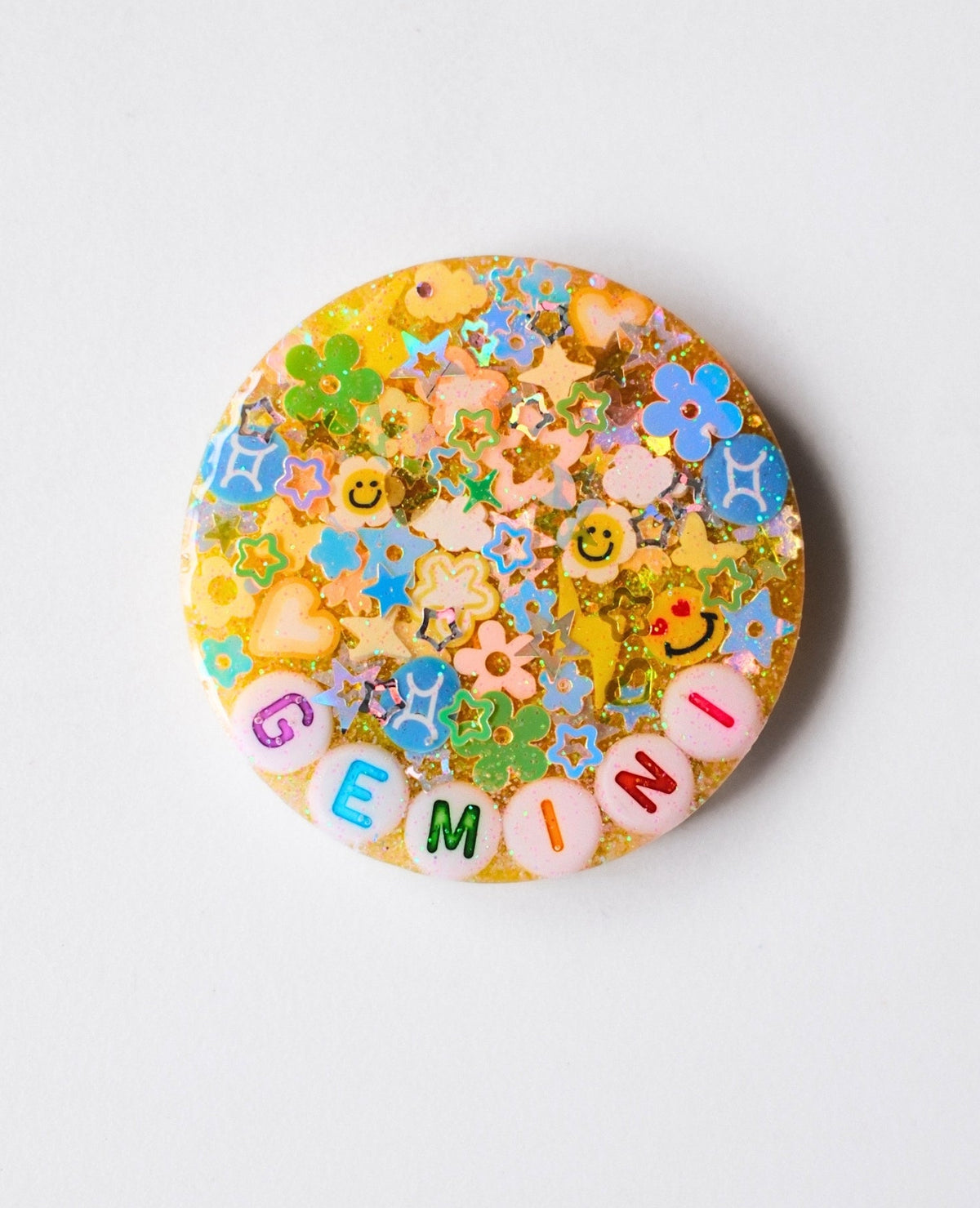 A round resin charm or button featuring a colorful Gemini zodiac design with glitter, small charms, and the letters 'GEMINI' at the bottom, shown on a light gray background.