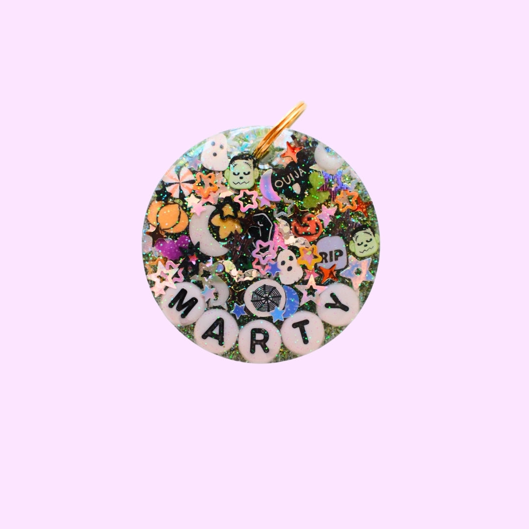 A round resin charm with a gold-tone loop, decorated with a colorful collage of Halloween-themed stickers, glitter, and white letter tiles spelling 'MARTY' on a light pink background.