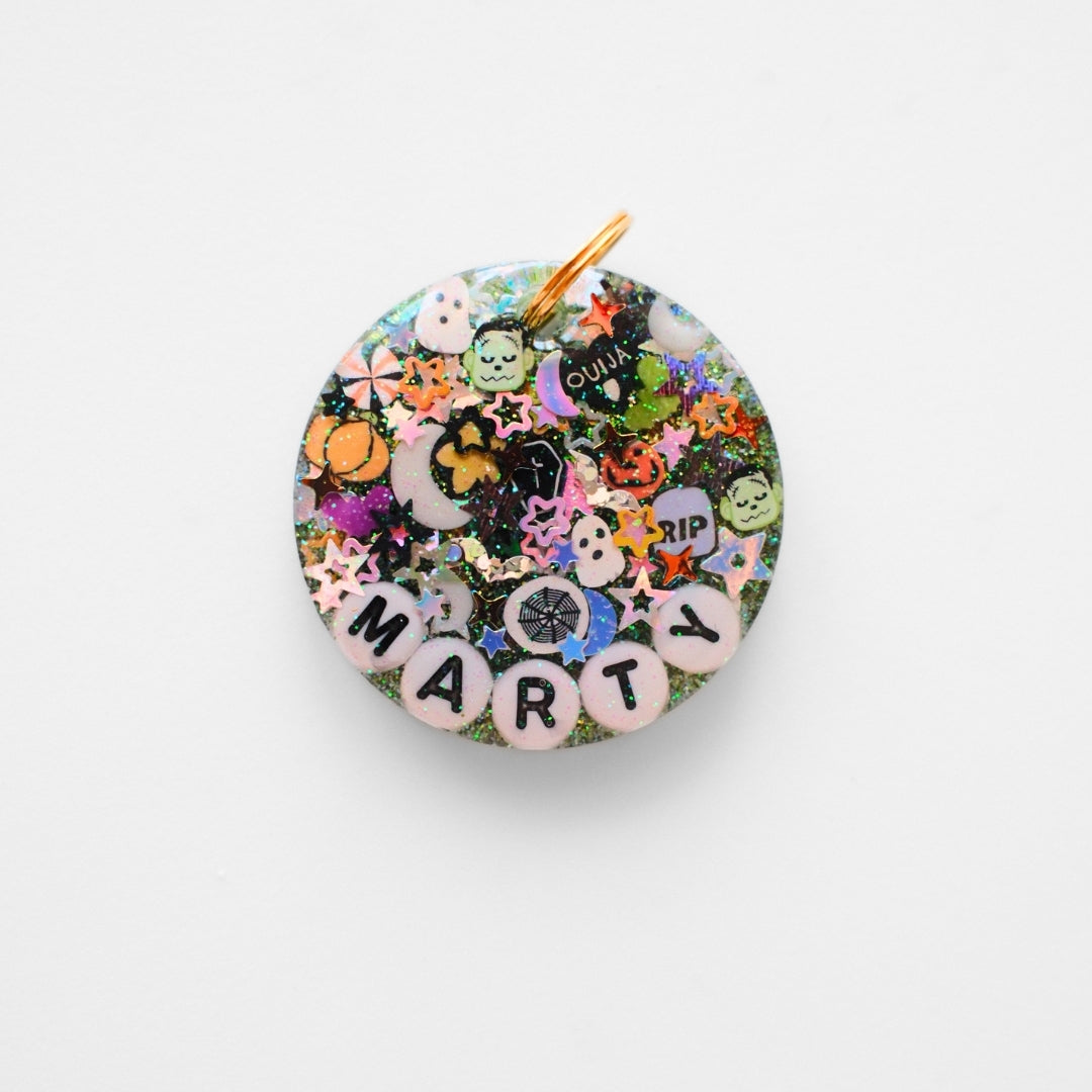 A round resin charm pendant featuring a colorful collage of Halloween-themed stickers, including ghosts, bats, a jack-o'-lantern, stars, and the word 'MARTY' spelled out in letter beads at the bottom, with a gold hanging loop at the top, shown on a plain white background.