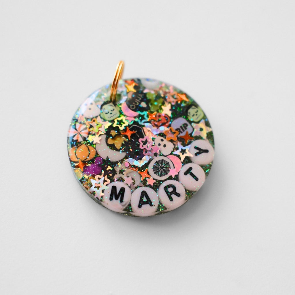 A round handmade charm with a gold loop at the top, filled with multicolor glitter, small star and flower pieces, and white letter beads spelling 'MARTY' on a light gray background.