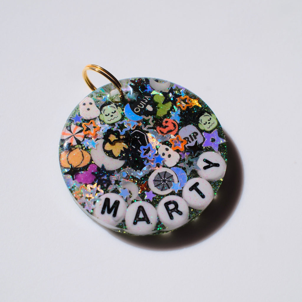 A round resin keepsake charm with a gold ring loop, filled with colorful Halloween-themed glitter, stickers, and white letter beads spelling 'MARTY' on a plain white background.