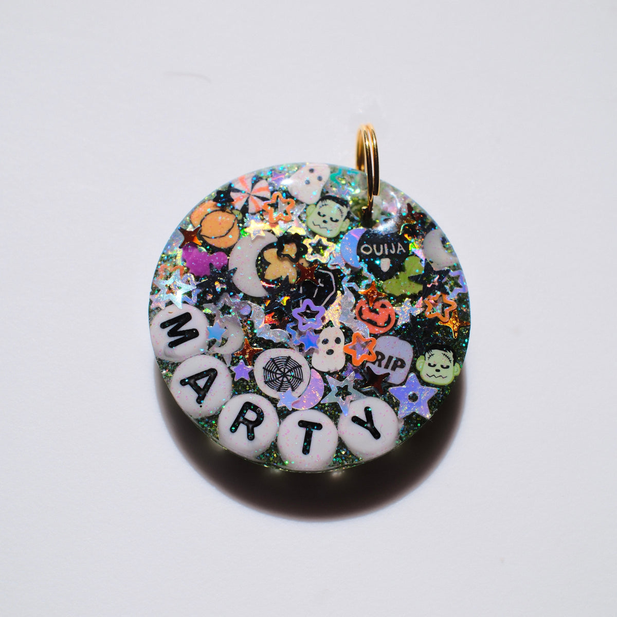 A round resin charm pendant with a gold-tone loop at the top, filled with colorful glitter, Halloween-themed stickers, and white letter beads spelling 'MARTY' on a plain white background.