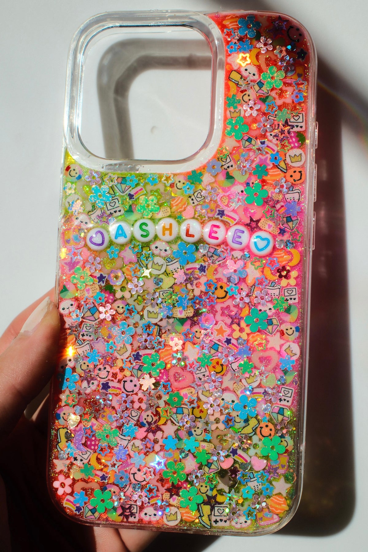 A clear phone case filled with colorful glitter, small flower charms, and decorative letter charms spelling 'JASHEEB', held in a hand against a plain light background.