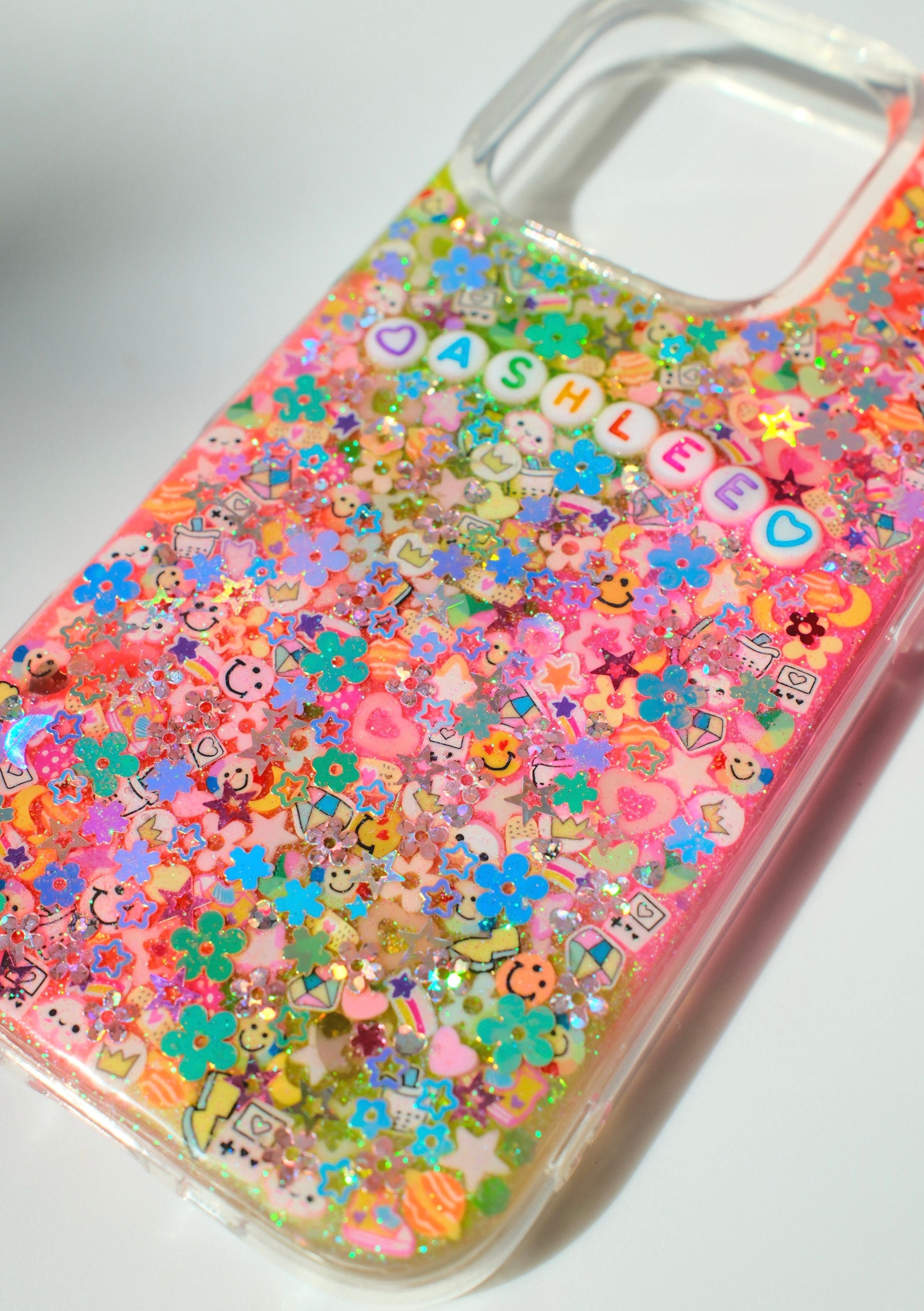 A clear phone case filled with dense, colorful glitter flakes and small decorative charms, including flowers, hearts, stars, and letter tiles spelling 'ASHLEEB'.