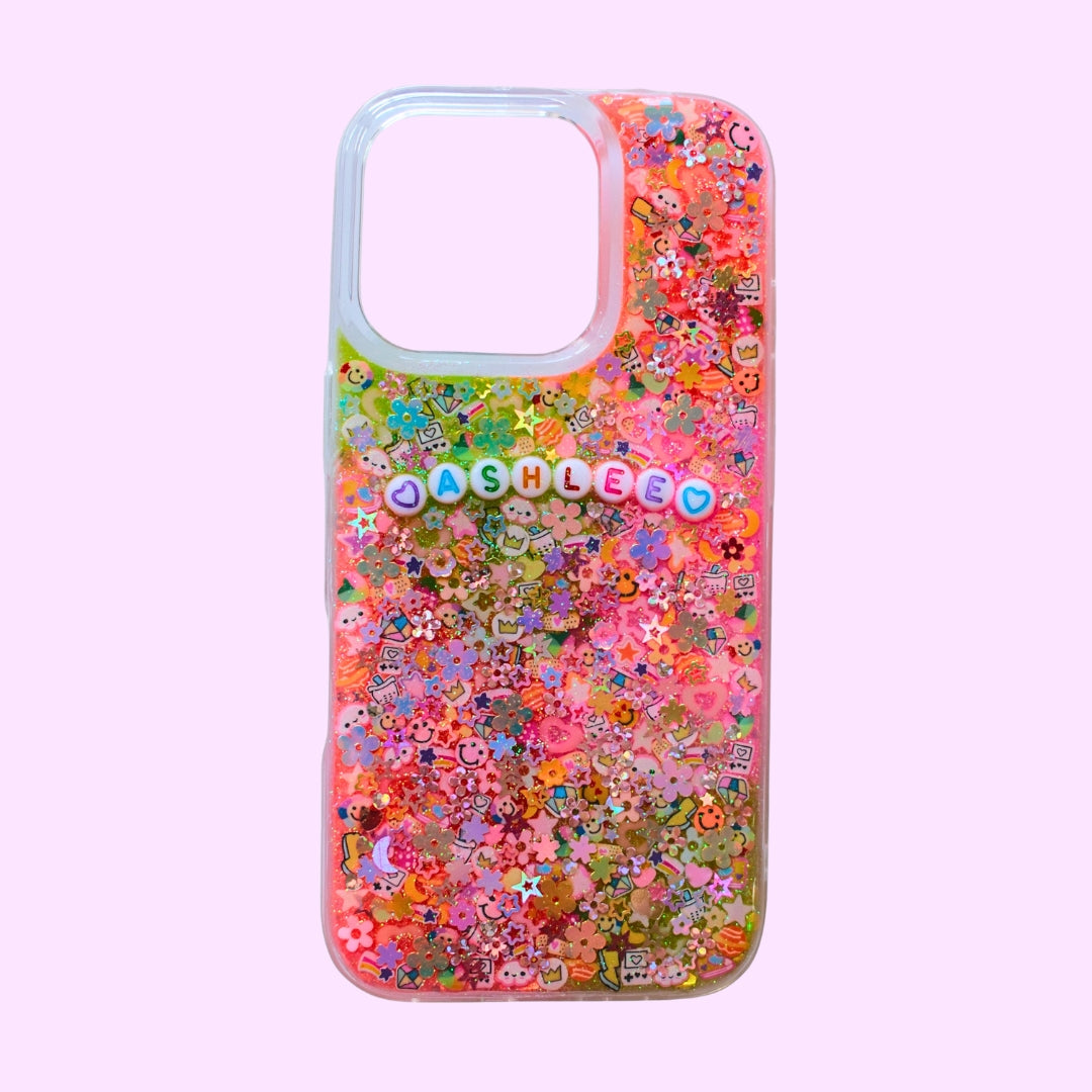 A clear phone case filled with colorful glitter and small decorative charms, including a prominent 'ASHLEEO' name charm, shown against a solid light pink background.