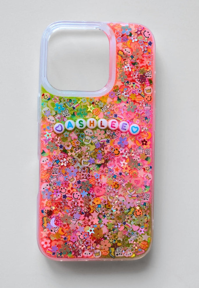 A clear iPhone case filled with colorful floral and cartoon glitter, featuring the name 'CASHLEE' in pastel letter beads across the center, shown on a neutral gray background.