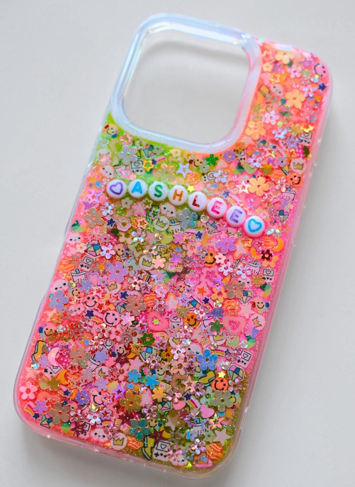 A clear phone case filled with colorful glitter flakes and small charms, including flowers and the name 'CASHLEE' in raised letter beads, shown on a light gray background.