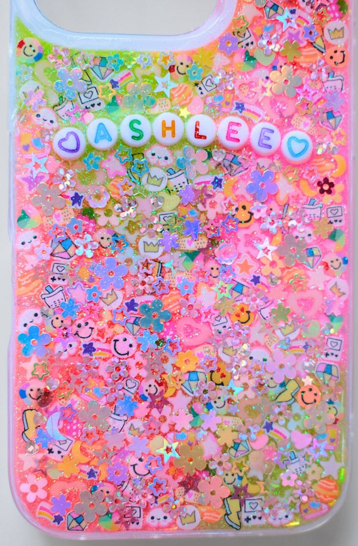A clear phone case filled with dense pink glitter, small pastel charms, and decorative elements, featuring the name 'JASHLEE' in pastel letter charms across the upper portion.