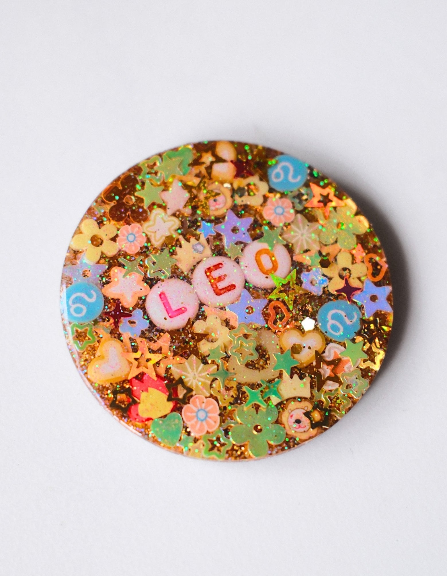 A round pinback button featuring a colorful, glittery astrological design for Leo, with the letters 'LEO' spelled out in pink tiles and surrounded by stars, flowers, and the number 6, set against a plain light gray background.