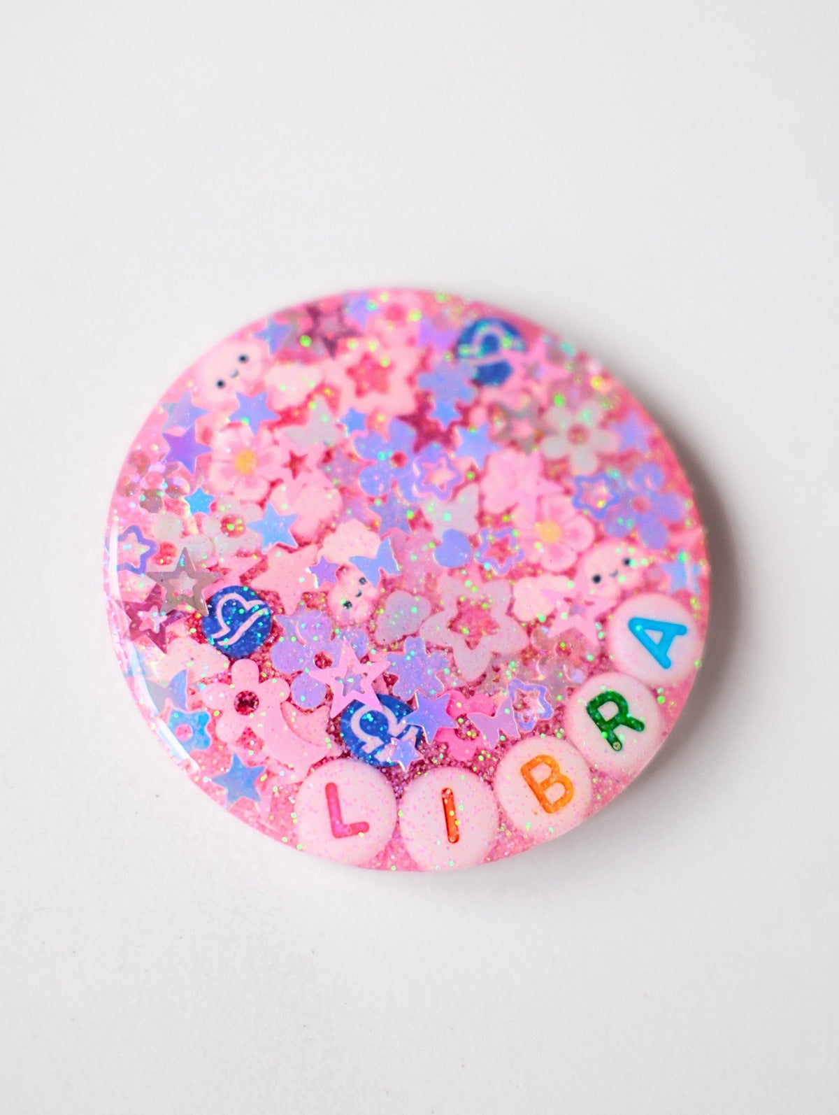 A round pink glitter pinback button featuring the word 'LIBRA' spelled out in colorful letter beads, surrounded by pastel glitter stars, flowers, and small charms, shown on a light gray background.