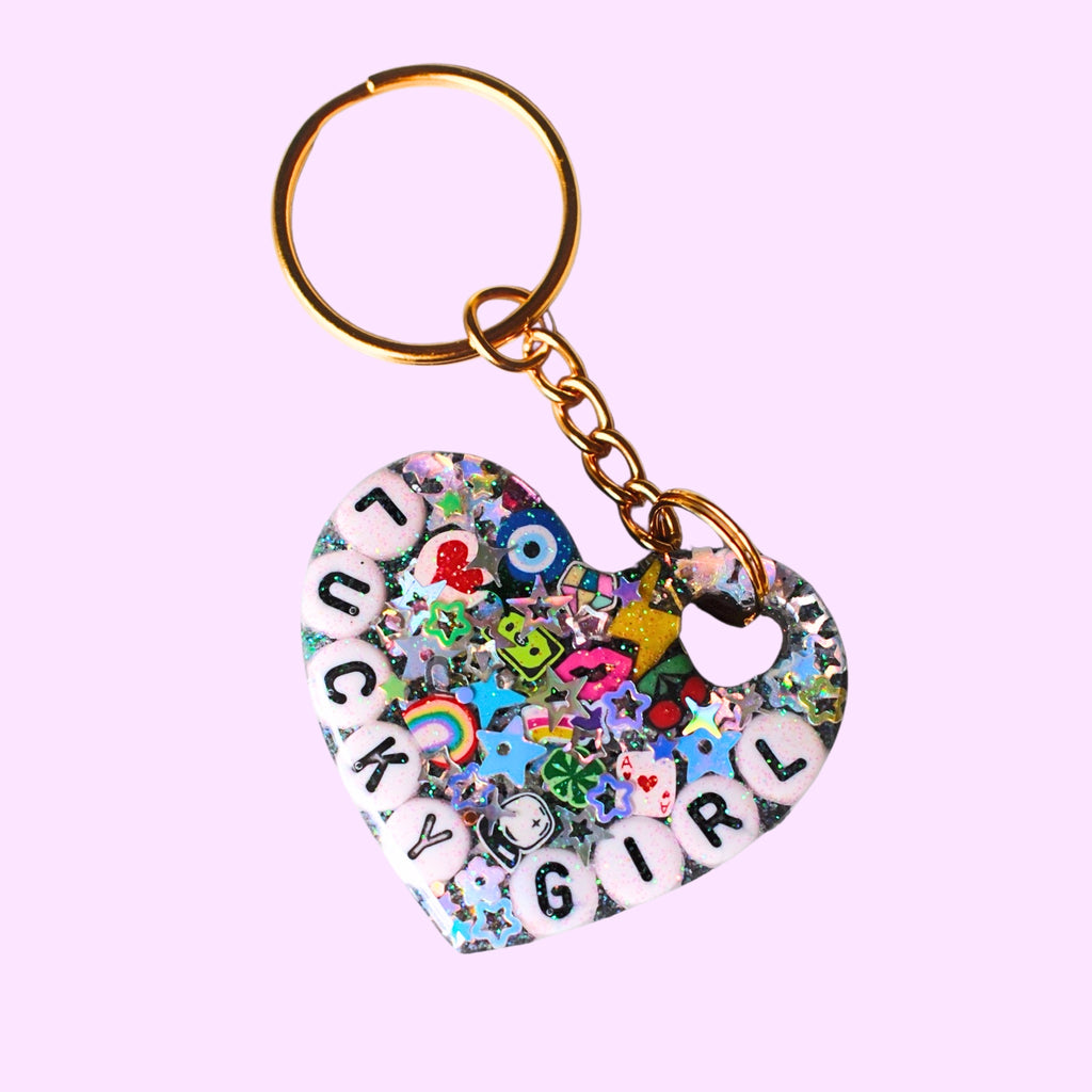 A heart-shaped keychain charm filled with colorful glitter, small charms, and white letter tiles spelling 'LUCKY GIRL', attached to a gold-tone split ring and chain on a light gray background.