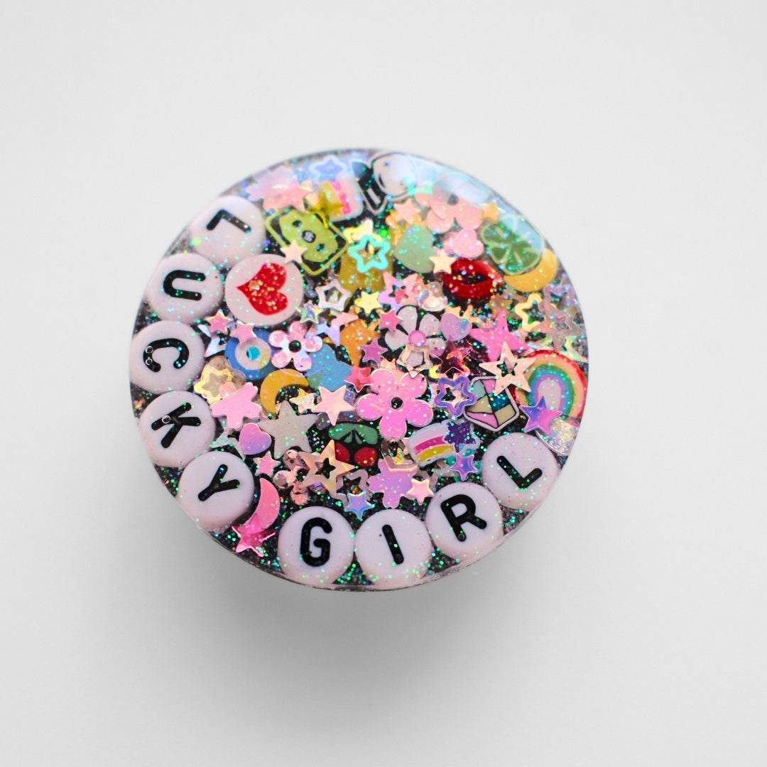 A round, flat-lay product shot of a shaker lid or button featuring a colorful 'LUCKY GIRL' design. The design includes white letter beads spelling 'LUCKY GIRL' on the outer edge, surrounded by a dense mix of pink, green, yellow, and blue glitter, flowers, stars, and small charms on a light gray background.