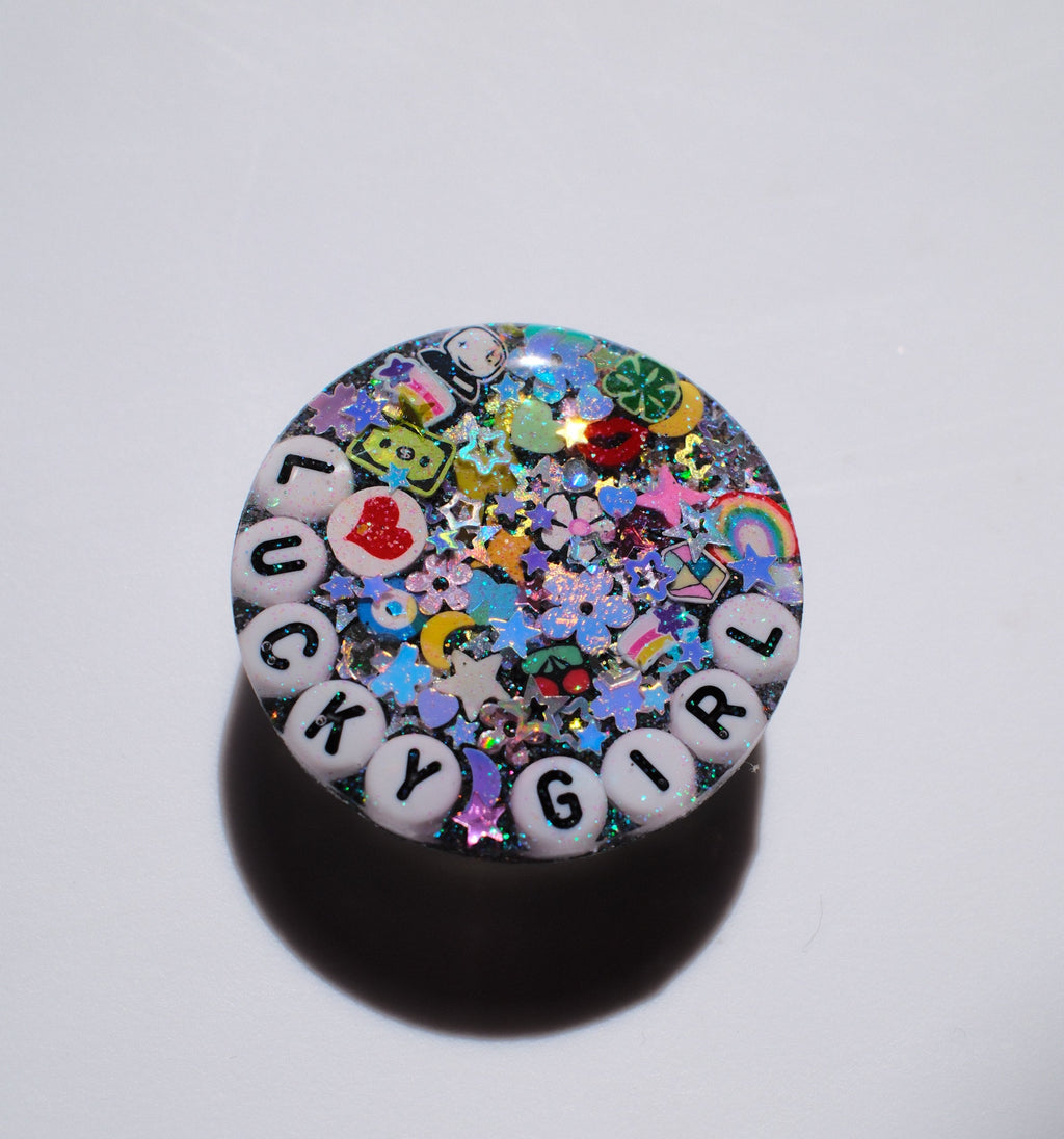 A round, glossy decorative item featuring the phrase 'LUCKY GIRL' spelled out by white letter beads, surrounded by a dense, colorful array of glitter, charms, and confetti. The product is photographed on a plain white surface with a soft shadow underneath.