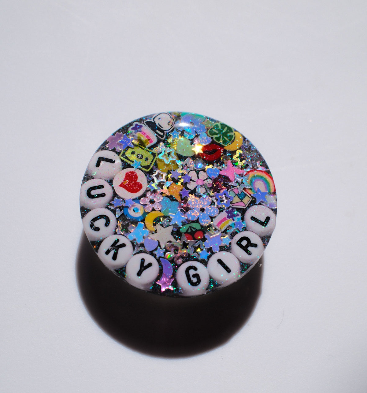 A round, glossy decorative item featuring the phrase 'LUCKY GIRL' spelled out by white letter beads, surrounded by a dense, colorful array of glitter, charms, and confetti. The product is photographed on a plain white surface with a soft shadow underneath.