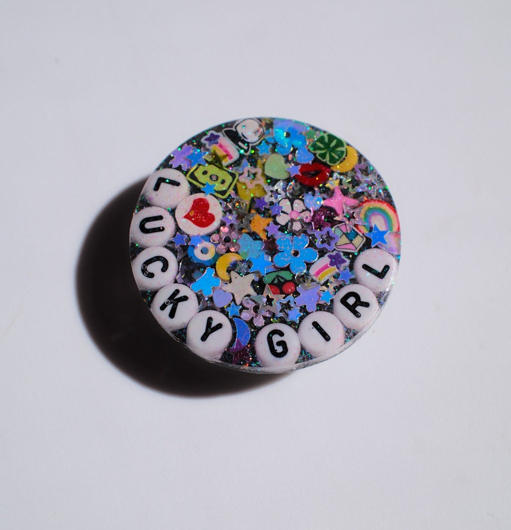 A round, multicolor glittery charm with white letter beads spelling 'LUCKY GIRL' on a plain white background.