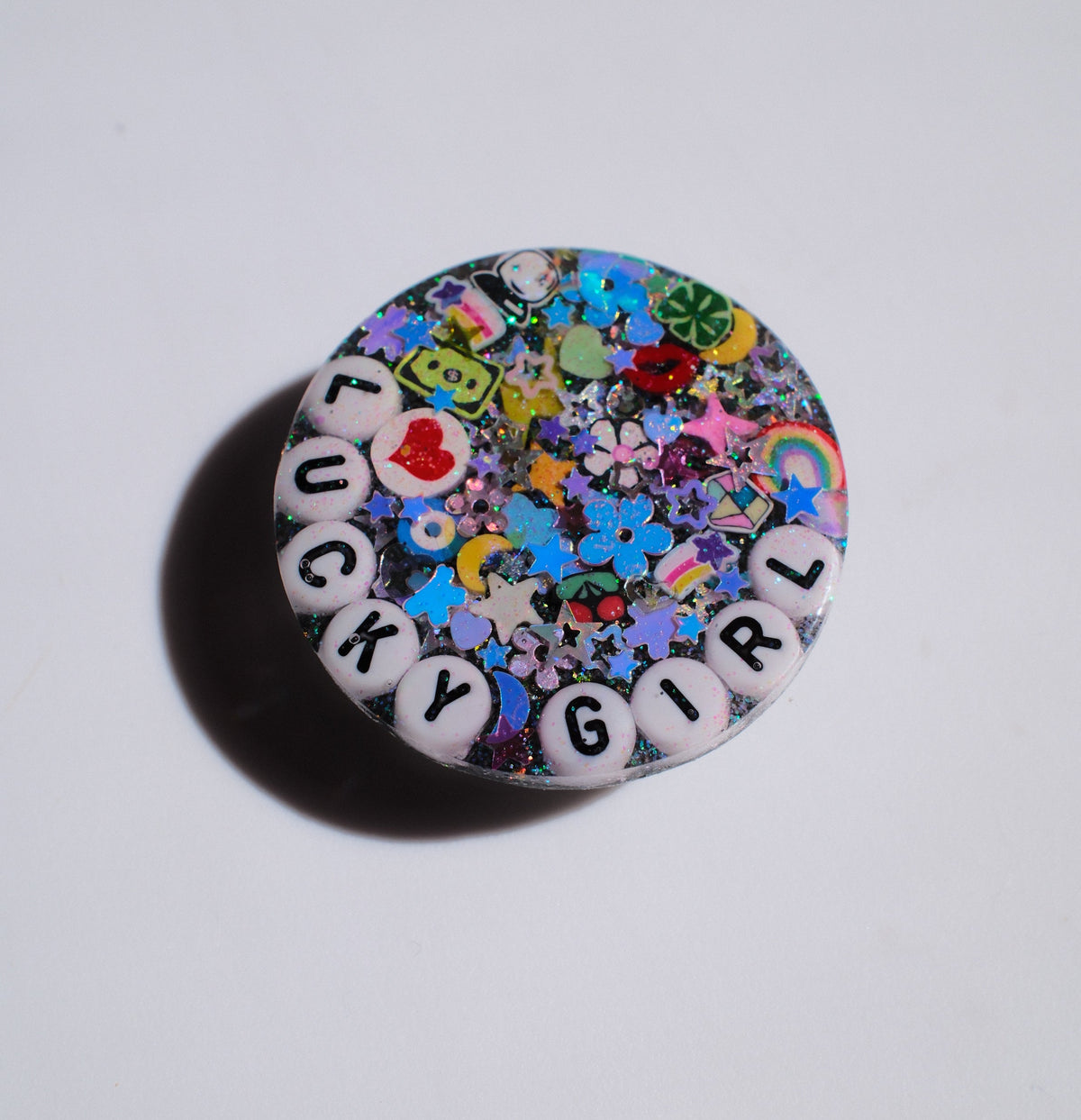 A round, multicolor glittery charm with white letter beads spelling 'LUCKY GIRL' on a plain white background.