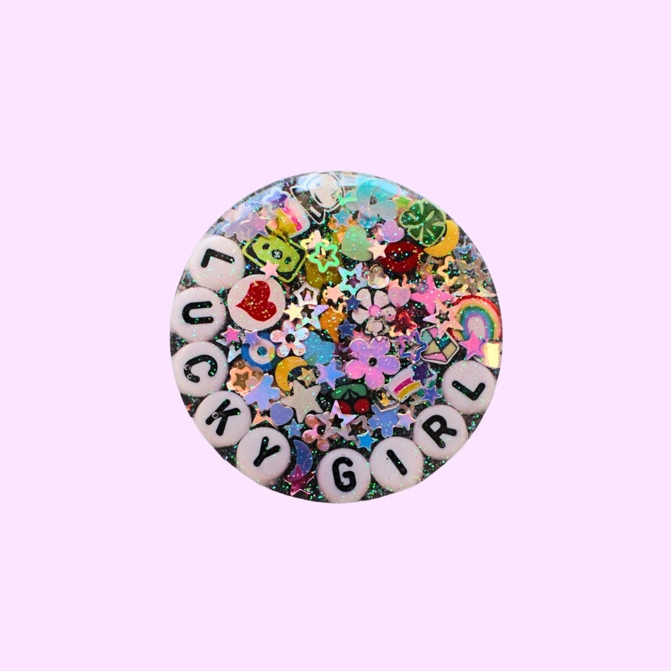 A round pinback button featuring a colorful, glittery 'Lucky Girl' design with white letter beads spelling out the words and a multicolor confetti background, set against a solid light purple background.