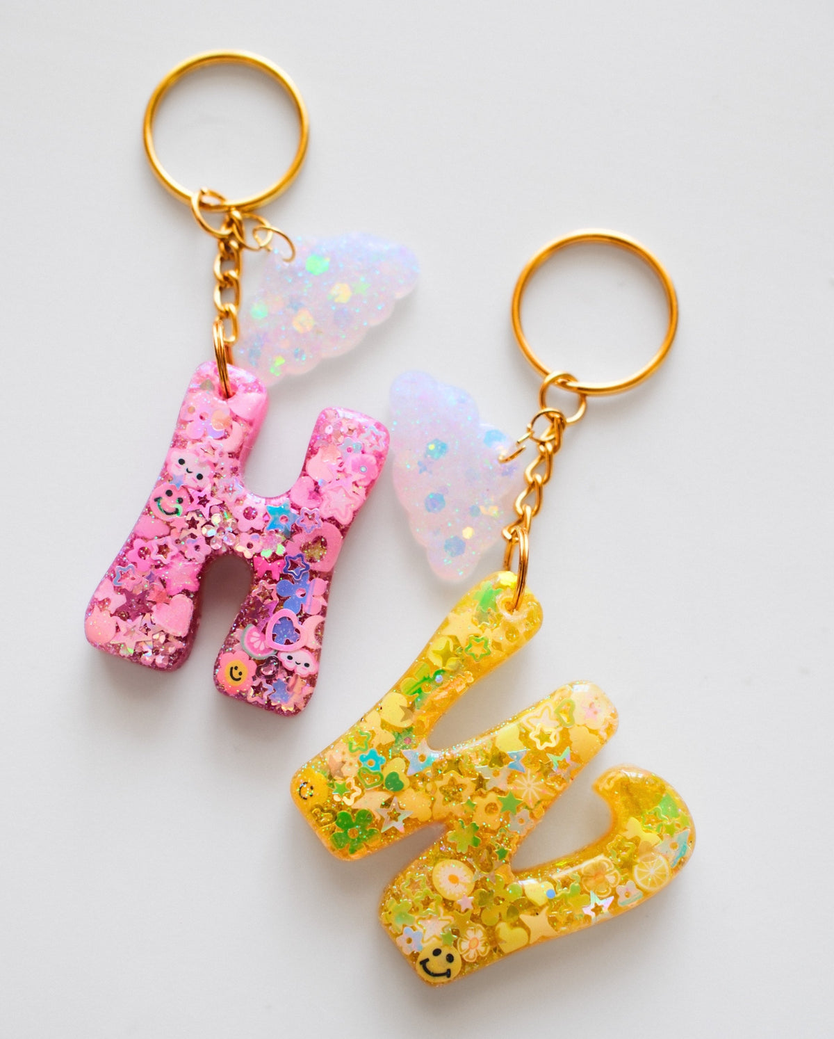 Two glittery resin letter keychains spelling 'HM', one in pink and one in yellow, each with a gold key ring and chain, shown on a light gray background.