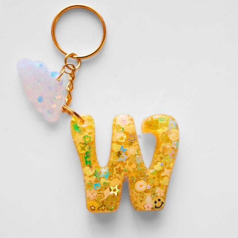A gold metal keychain featuring a large, glittery yellow resin letter 'W' filled with colorful confetti and small star charms, accompanied by a small iridescent white cloud charm, shown on a light gray background.