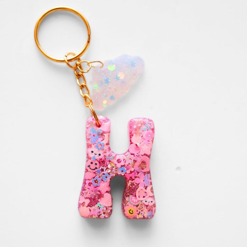 A pink glittery letter 'H' keychain with a gold-tone split ring and chain, featuring a small iridescent heart charm, on a light gray background.