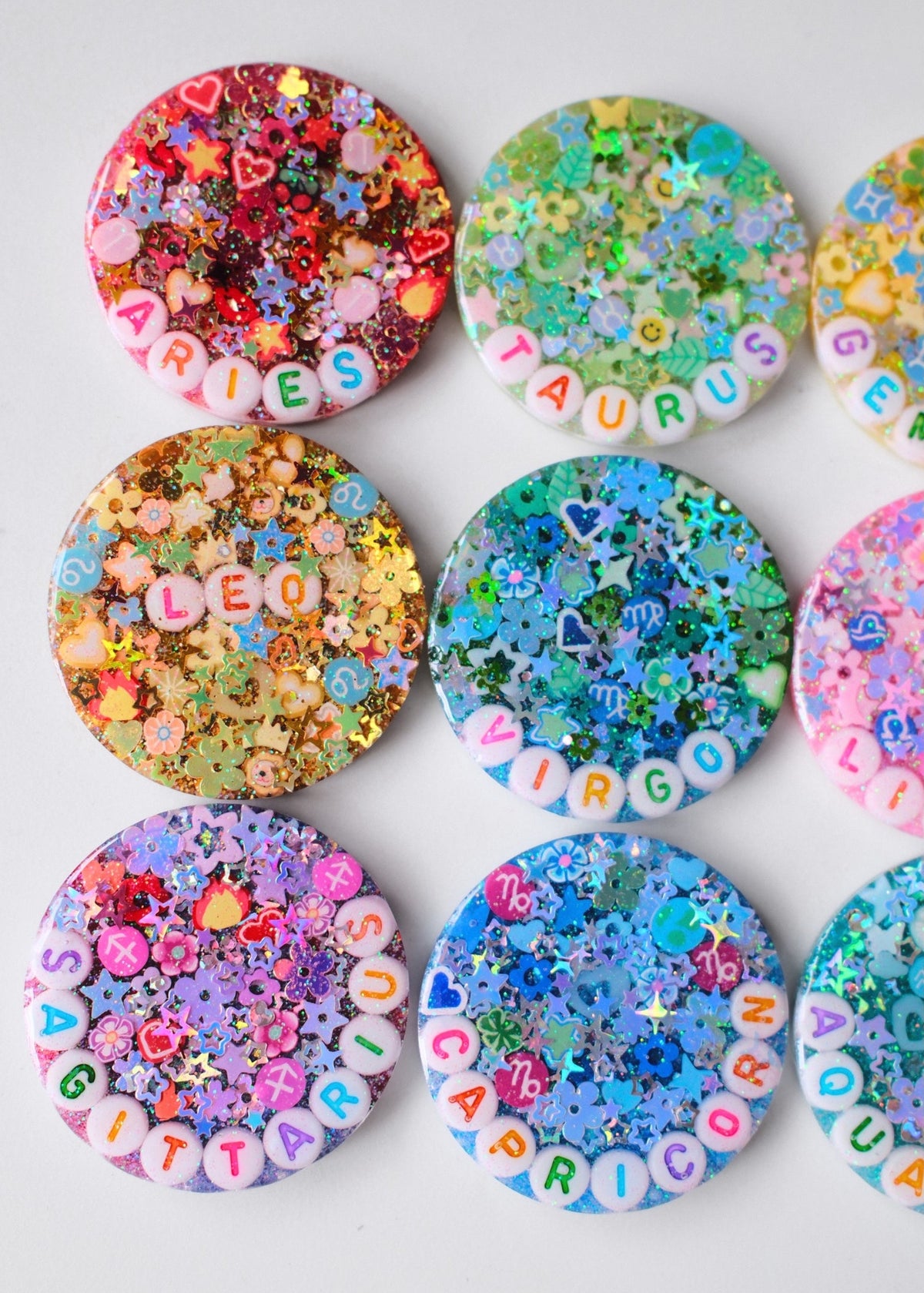 Nine round glittery resin pendants arranged in a 3x3 grid on a light gray background. Each pendant features colorful flower, heart, and star glitter, with some displaying letter beads spelling out names like 'MARTHA', 'LEO', 'ZIRGO', 'PANDORA', and 'BITTER'.