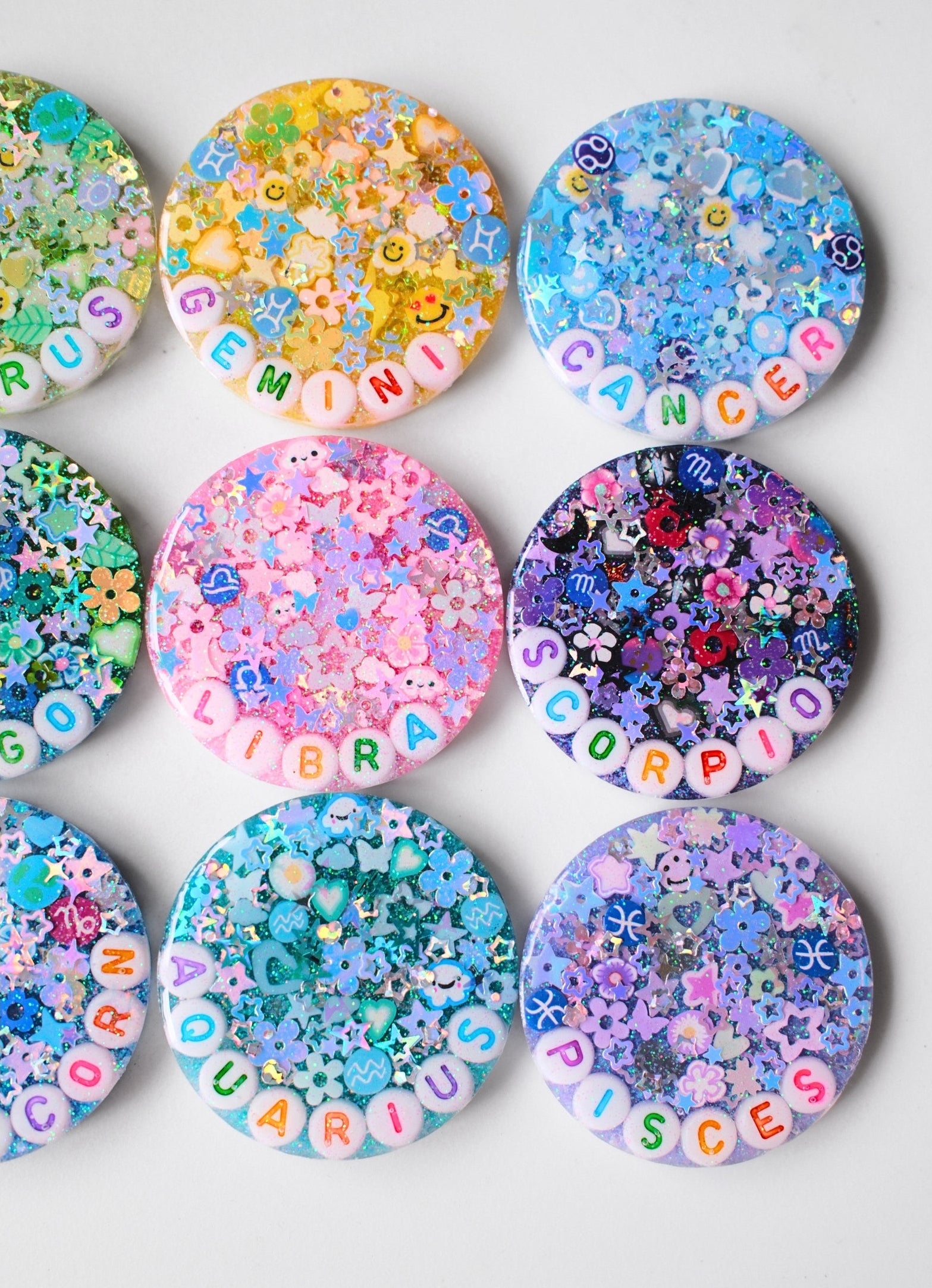 Nine round resin coasters arranged in a 3x3 grid on a light gray background. Each coaster features a different zodiac sign name spelled out in colorful letter tiles, with dense multicolor glitter, flower charms, and beads filling the top portion.