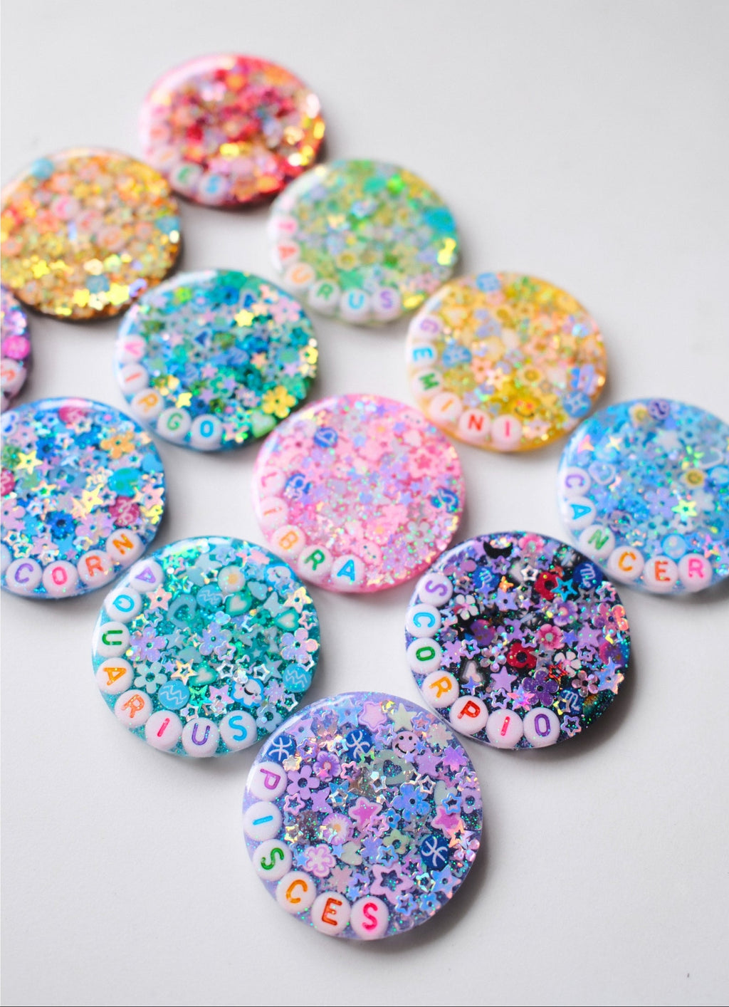 A group of round glittery resin buttons or charms arranged on a light gray background. Each button features colorful floral glitter, small letter beads spelling out words, and pastel lettering, with some buttons showing holographic glitter.