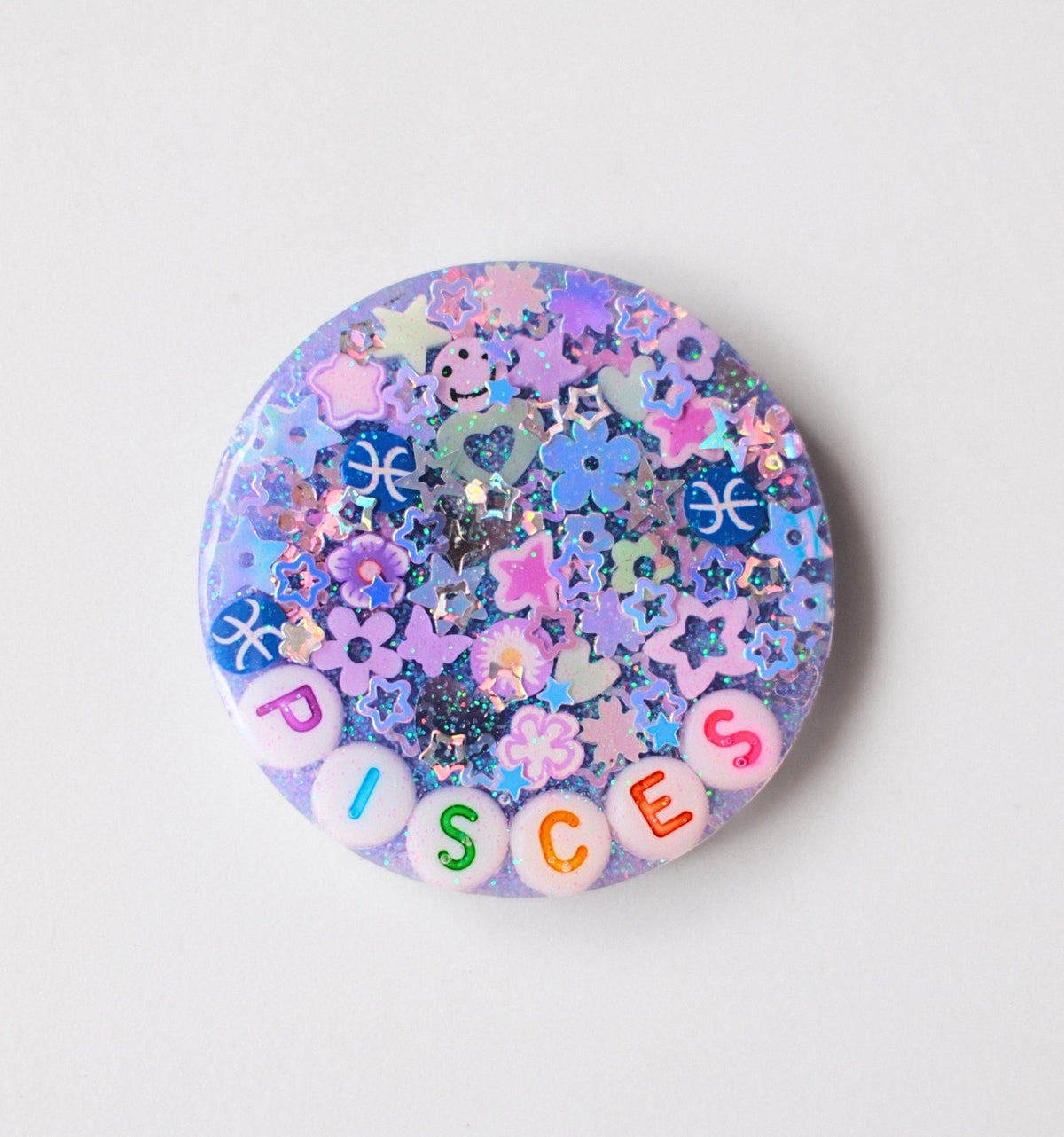 A round pinback button featuring a glittery lavender and blue background filled with colorful flower and star-shaped confetti, with the word 'DISCES' spelled out by pastel letter beads along the bottom edge.