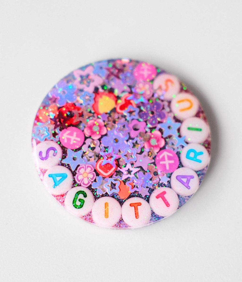 A round glittery pinback button featuring the word 'SUGAR' spelled out in colorful letter beads, surrounded by pink flowers, small hearts, and purple glitter.