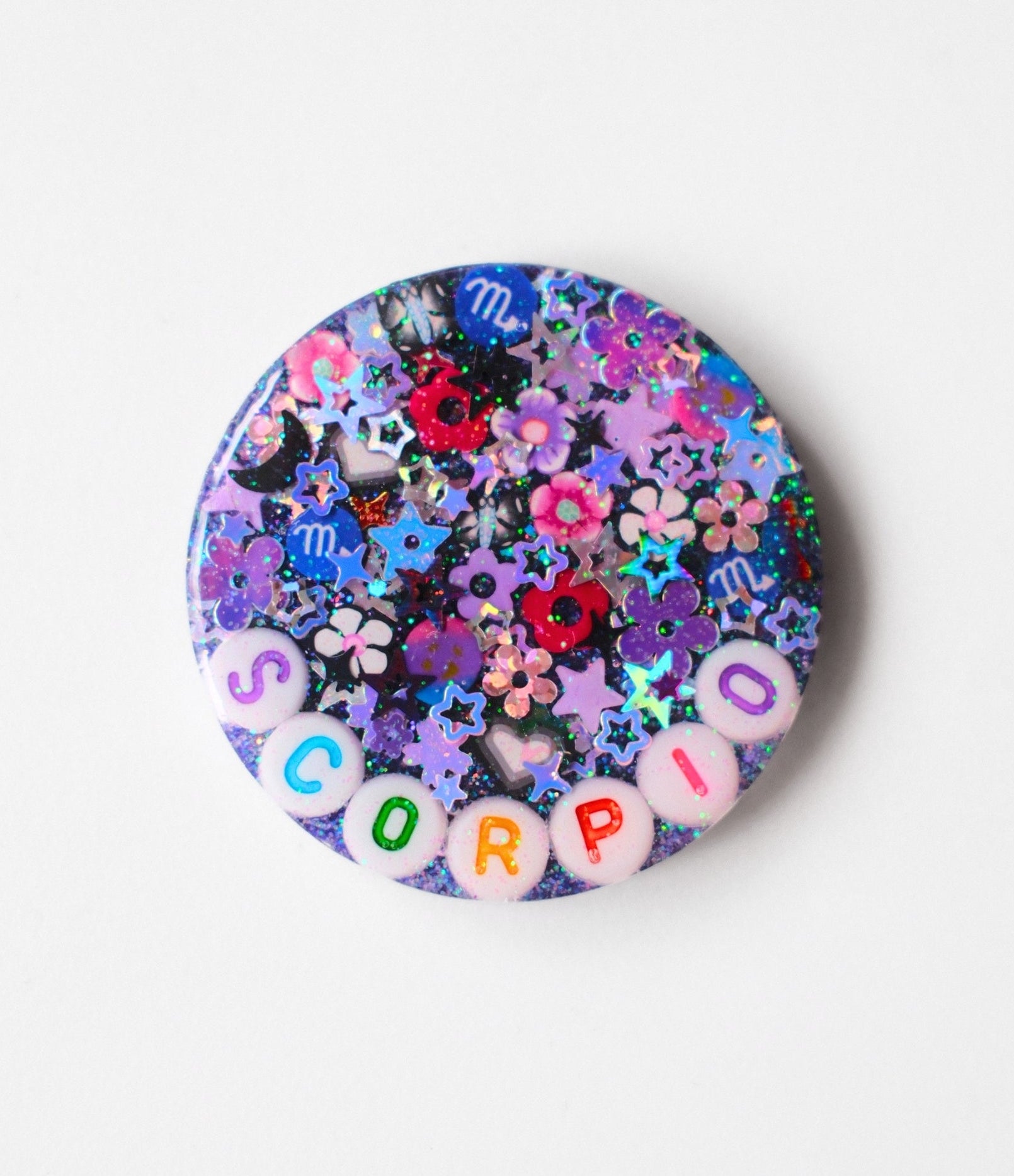 A round pinback button featuring a colorful, glittery Scorpio-themed design with floral elements and the word 'SCORPIO' spelled out in letter beads along the bottom, set against a plain white background.