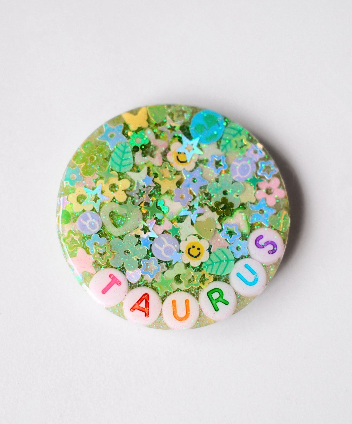 A round resin charm featuring a pastel green background filled with glitter, small flowers, leaves, and stars, with the word 'TAURUS' spelled out by colorful letter beads along the bottom edge.