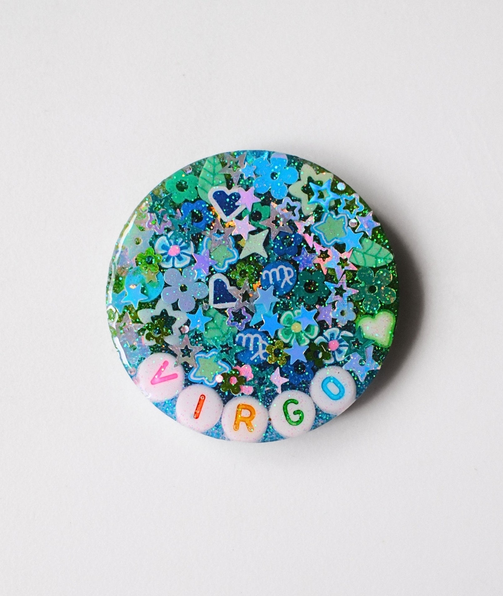 A round resin pinback button featuring a blue and green glittery background filled with star and heart charms, and the word 'VIRGO' spelled out in colorful letter beads along the bottom edge.