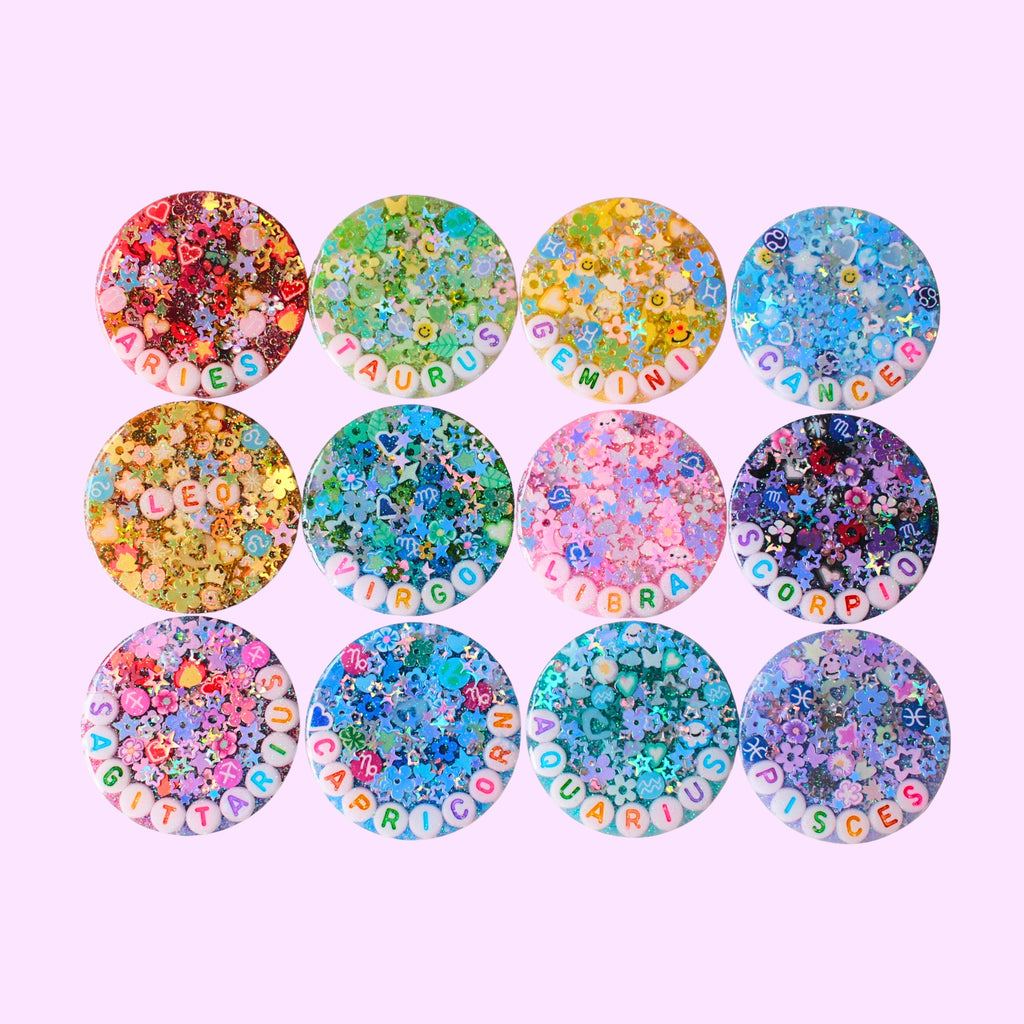 Twelve round resin charms arranged in a 3x4 grid on a light pink background. Each charm is filled with colorful glitter, small charms, and letter tiles spelling out the month name on the front.